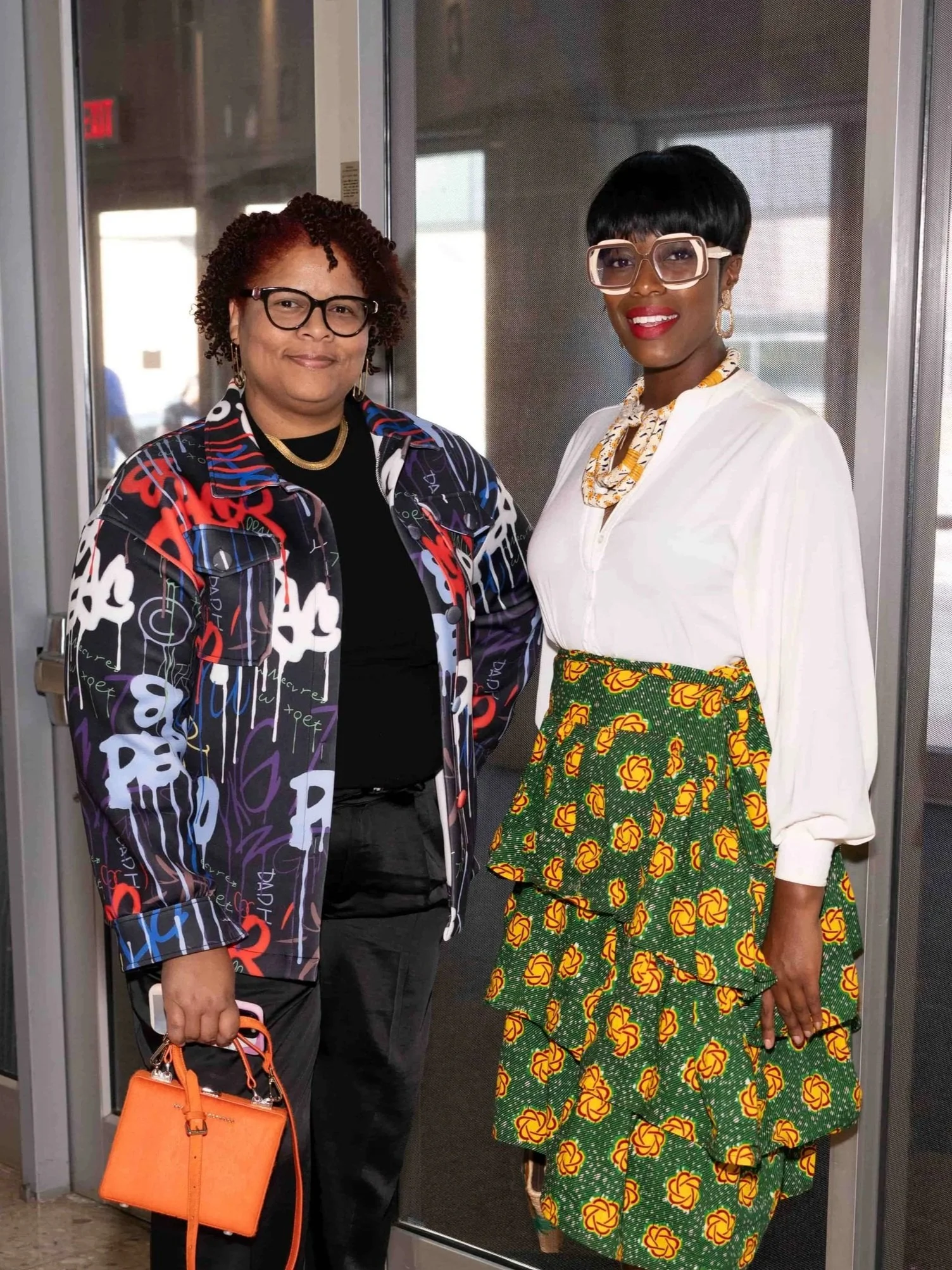 Two women standing together, one holding an orange purse, in an indoor setting near glass doors, smiling for the camera.