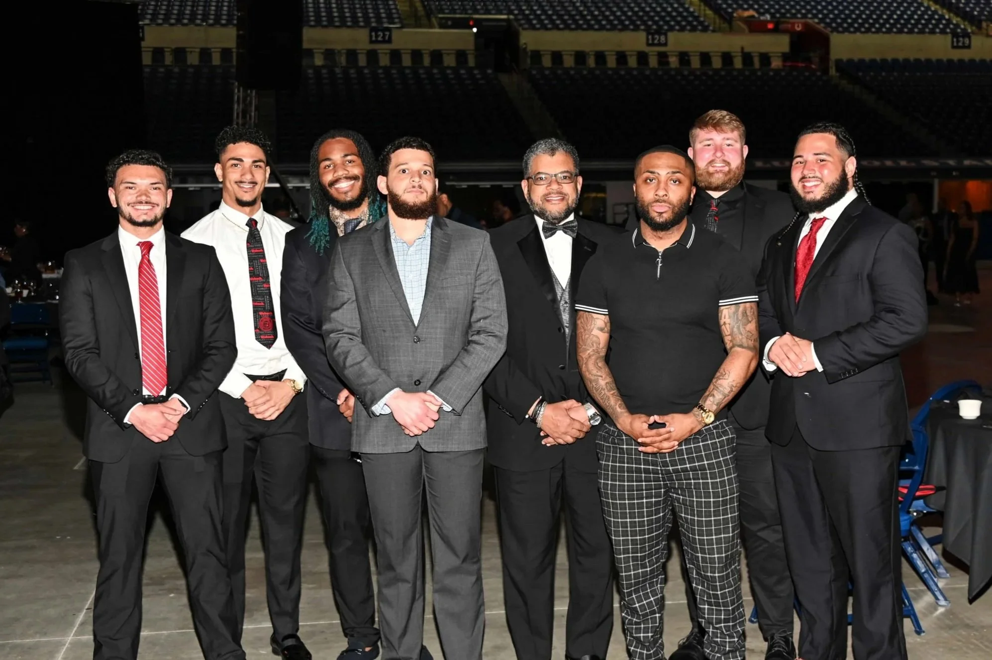 Group of nine men dressed in formal attire, standing together in an indoor event space. Most are wearing suits, and some have bow ties or ties. The background shows stadium seating and dim lighting.