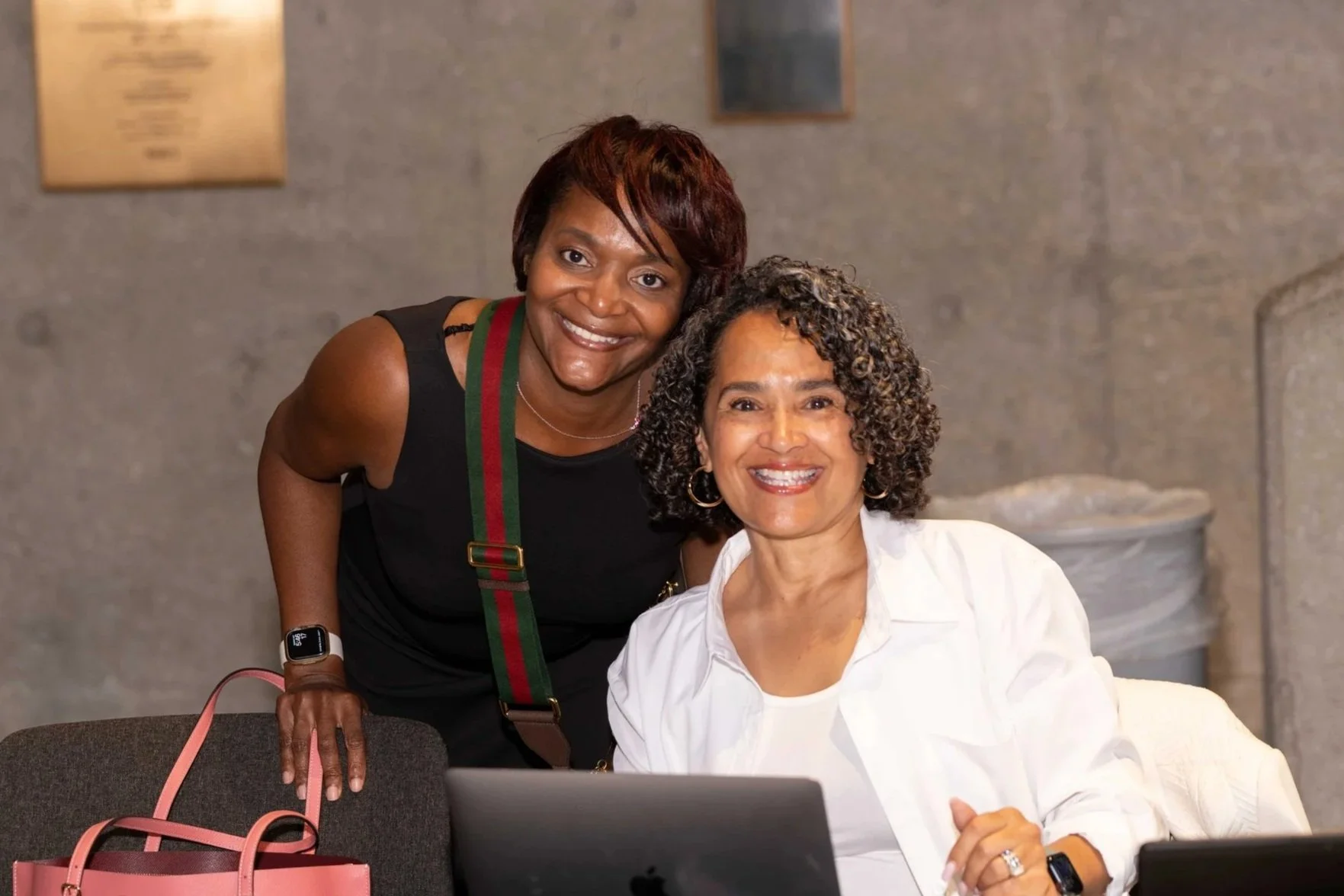 Two women smiling at a desk with laptops in an indoor setting.