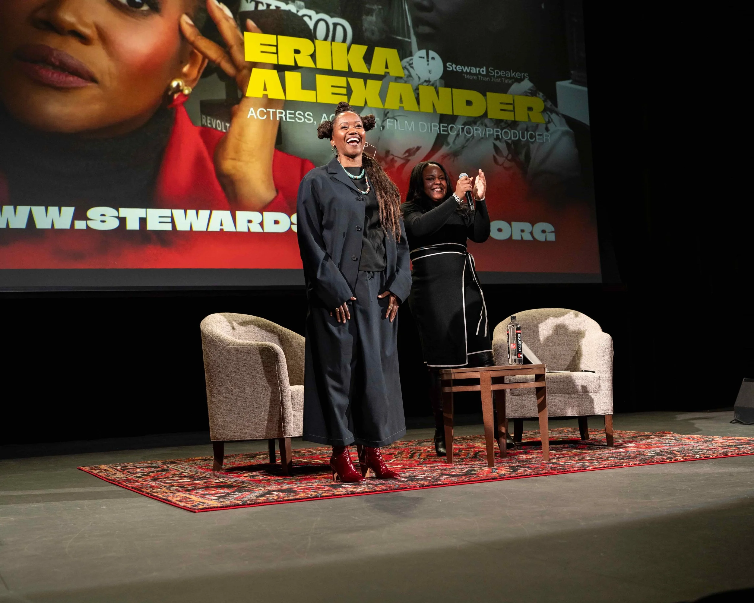 Two women on stage at a conference or event, with one woman smiling and the other holding a microphone. Behind them is a large screen displaying an image of a woman and text including "Erika Alexander" and a website link.