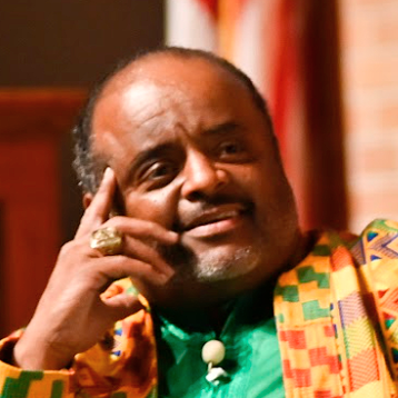 A man with a thoughtful expression, resting his head on his hand. He is wearing colorful clothing and a ring, with a blurred background that includes an American flag.
