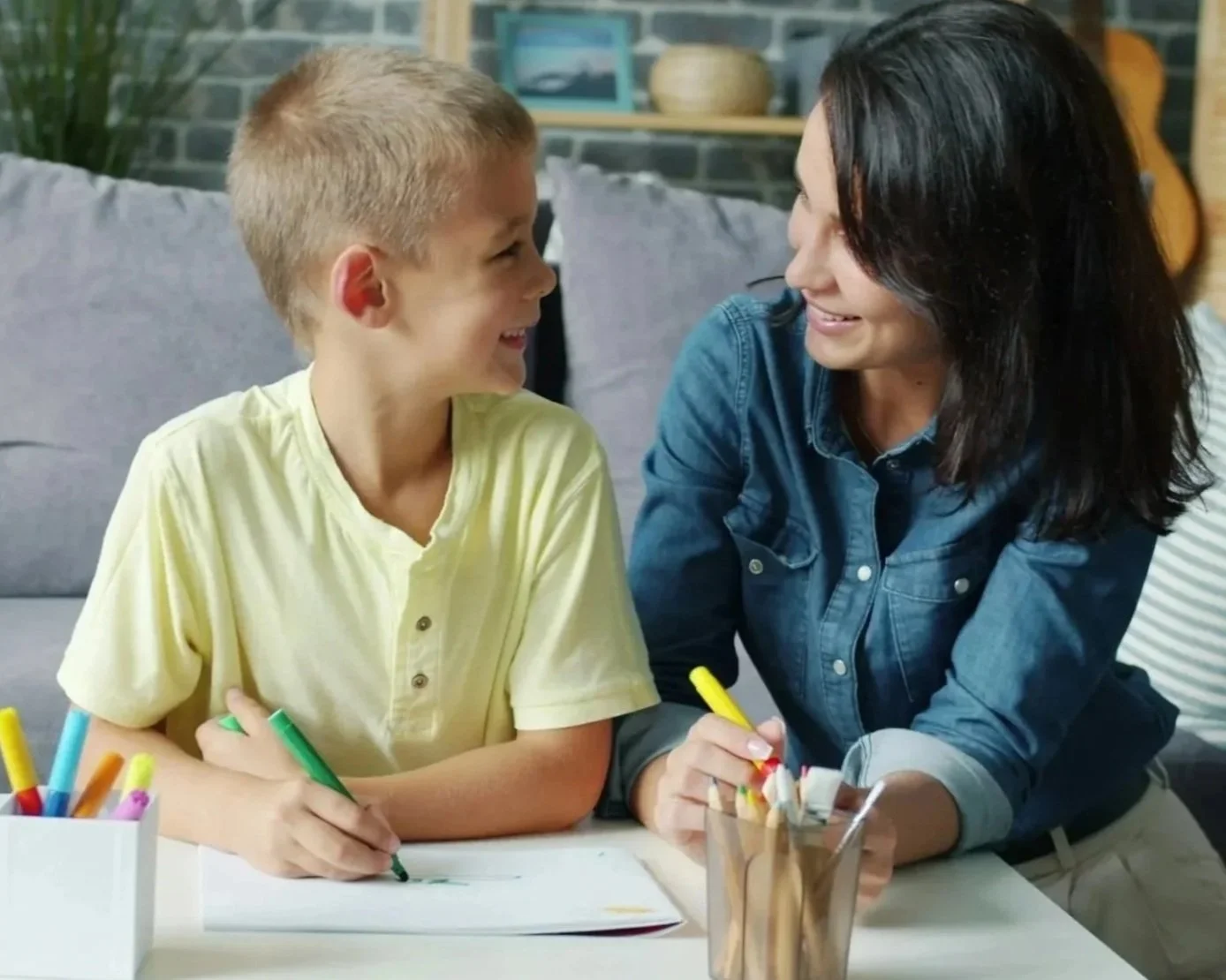 A young boy and a woman are sitting at a table, smiling at each other while drawing with markers. The boy is wearing a yellow shirt and the woman is wearing a blue denim shirt. They are in a cozy room with a brick wall and a guitar in the background.