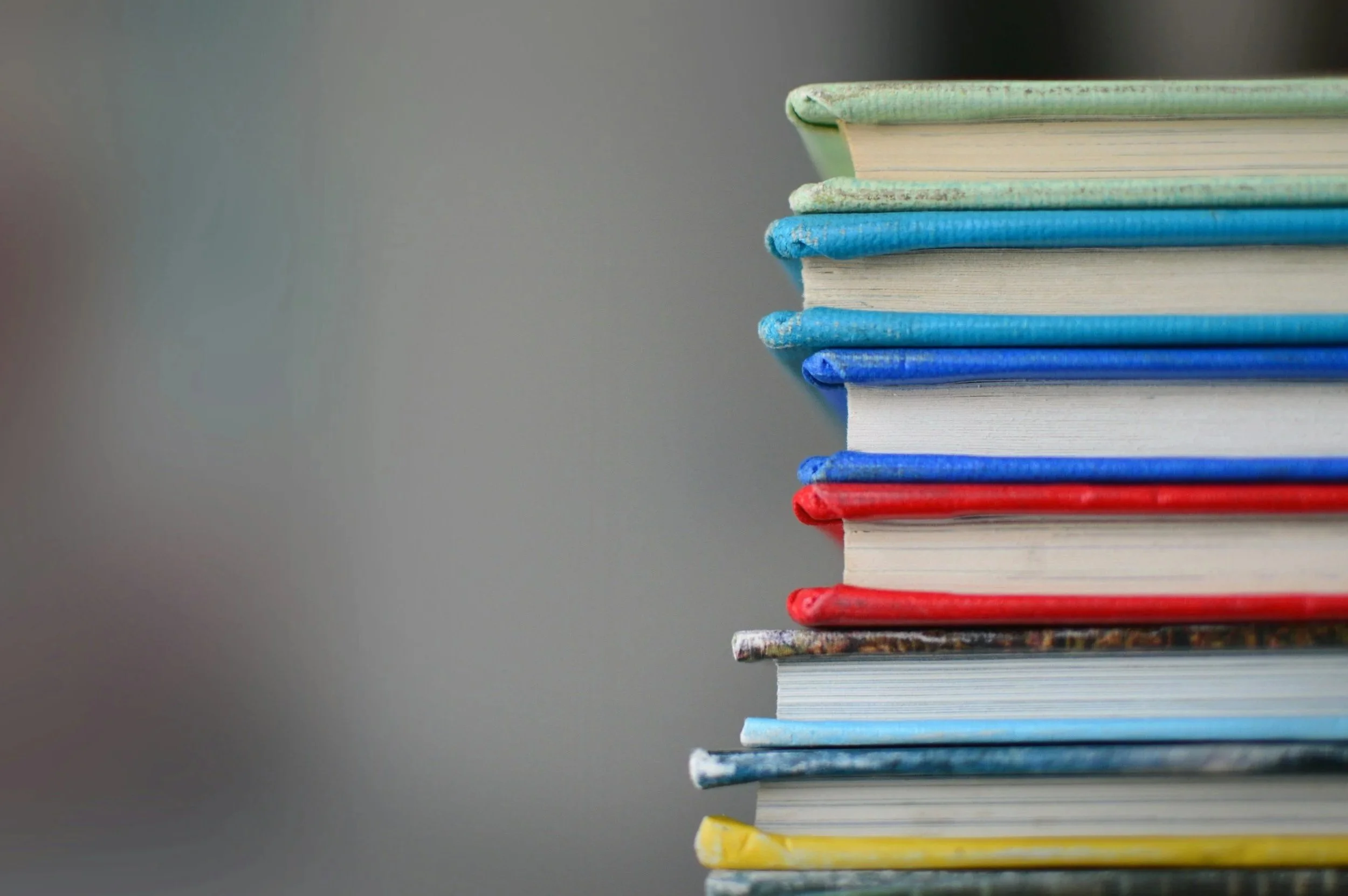 A stack of colorful books arranged vertically with bound covers, including shades of blue, green, red, and yellow, placed on a reflective surface against a plain, gradient background.