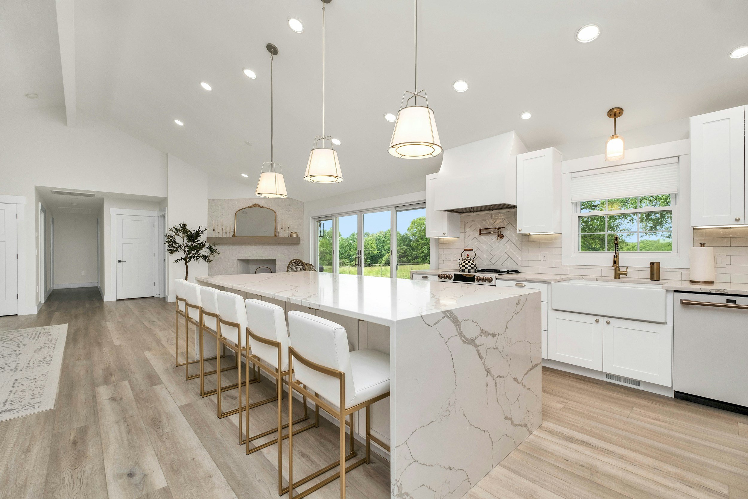 Modern white kitchen featuring a large marble island with six white and gold bar stools, white cabinetry, a window above the sink, and sliding glass doors leading to a green outdoor view.