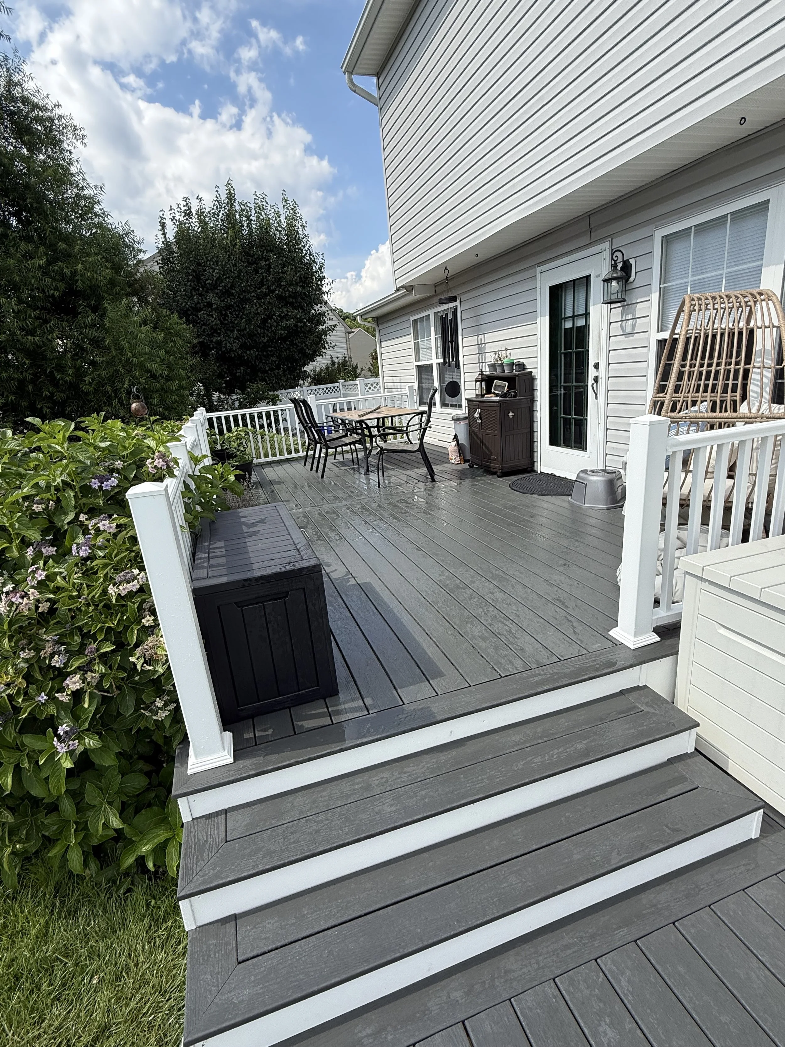 A backyard porch with a wooden deck, outdoor furniture, plants, and a house with white siding and a glass door, under a partly cloudy sky.