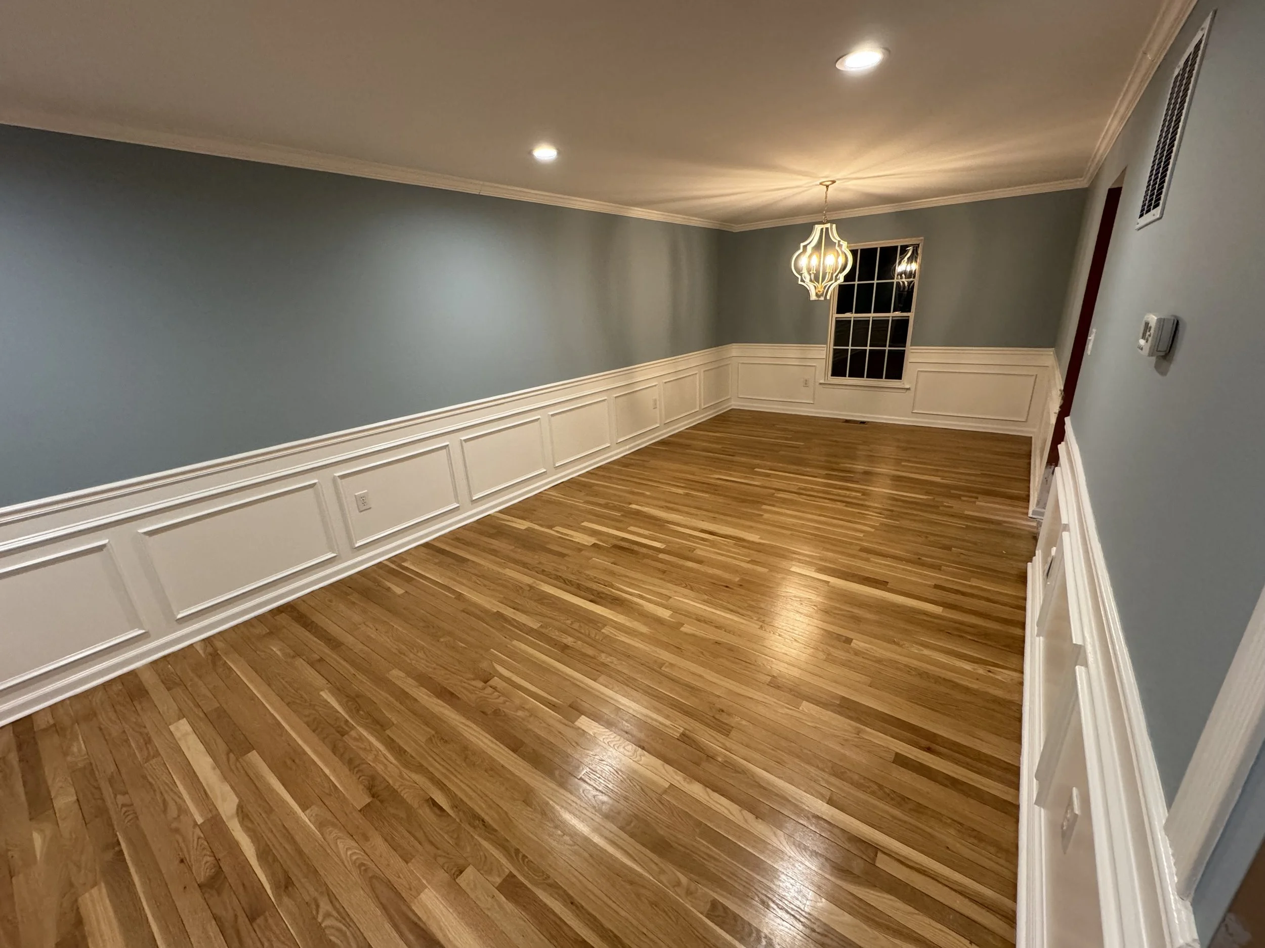Empty living room with hardwood floor, light blue walls, white wainscoting, ceiling lights, and a decorative pendant lamp near a window.