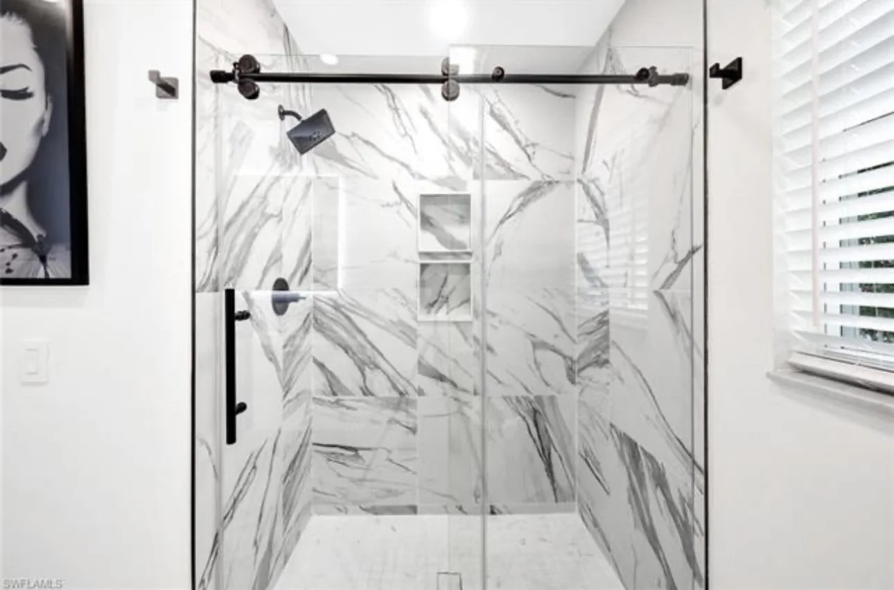 Modern walk-in shower with marble tiles, black metal hardware, a black showerhead, and a small built-in shelf, with white window blinds on the right.