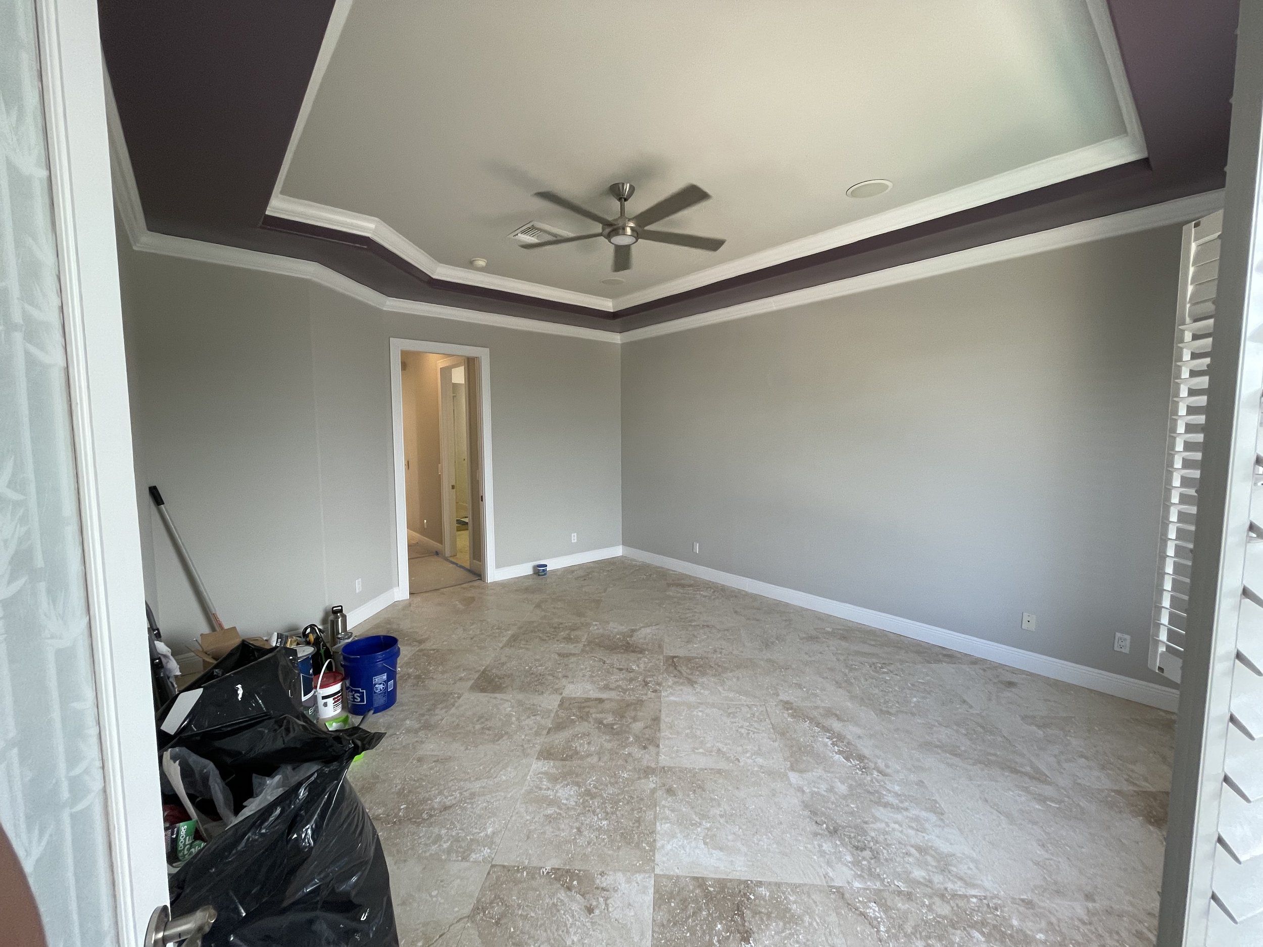 Empty living room with tiled floor, light gray walls, white trim, and a ceiling fan. Construction supplies and a trash bag are in the corner.