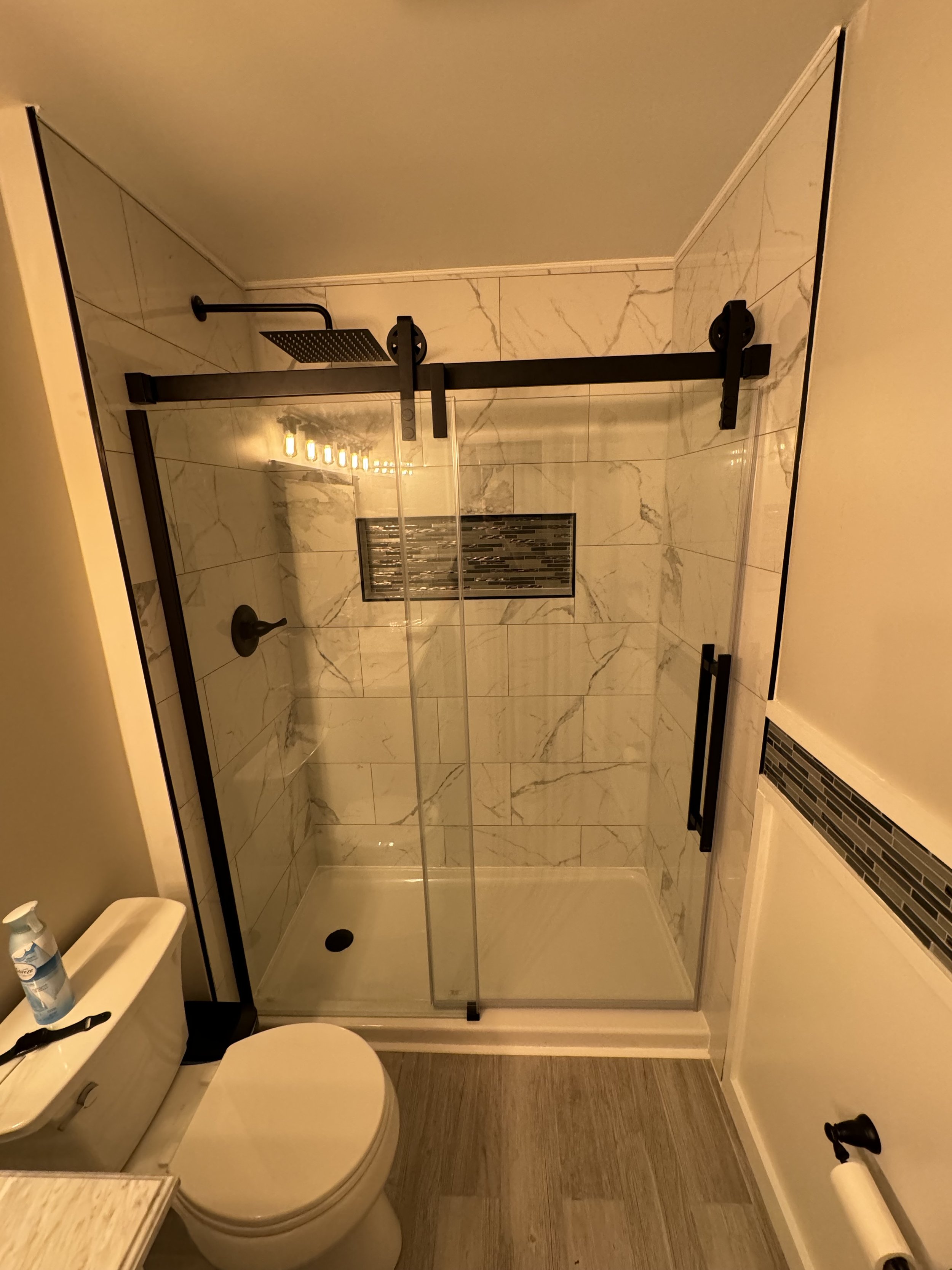 A bathroom with a walk-in shower featuring marble tiles, a black sliding barn door style shower door, a built-in tile niche for toiletries, and a wooden floor. The bathroom also includes a toilet with a bottle of cleaning spray and a black toothbrush