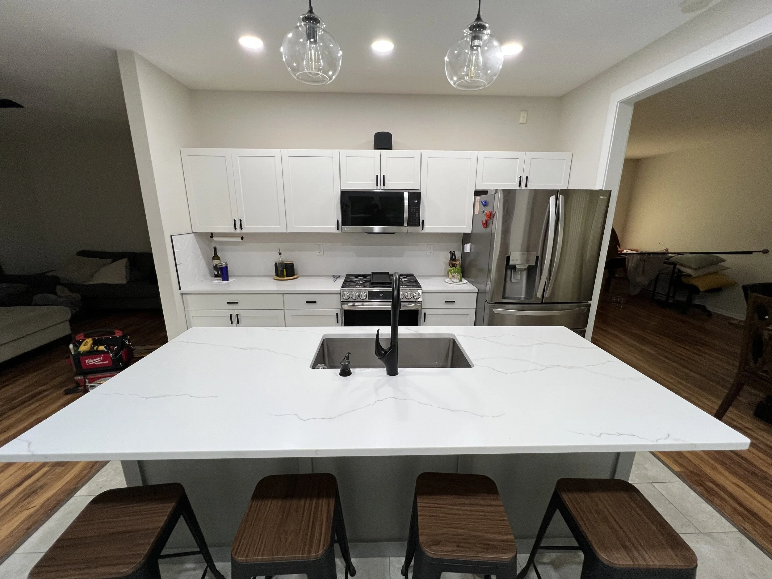 Modern kitchen with a white island countertop, stainless steel refrigerator, microwave, and oven, and white cabinets. There are bar stools around the island, and two pendant lights hang from the ceiling.