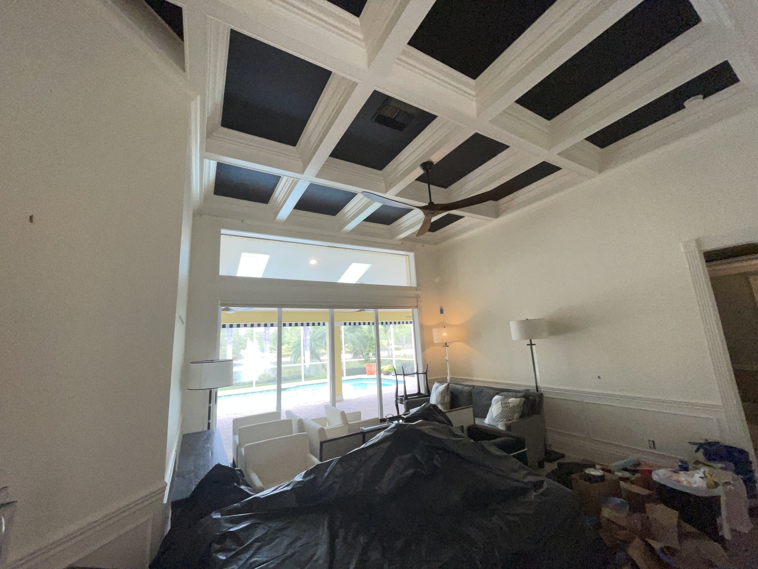 Living room with high coffered ceiling, ceiling fan, large sliding glass door leading outside to a pool area, corner lamps, sofa, chairs, and boxes and items on the floor.