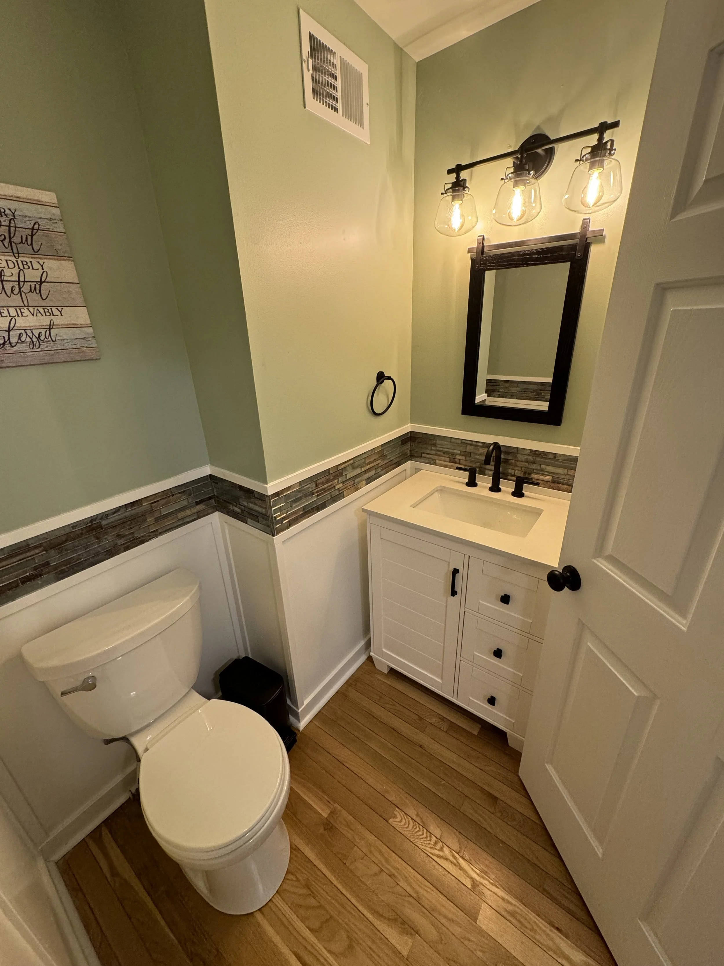 Small bathroom with light green walls, a white toilet, a white vanity with a black faucet, a framed mirror, and a three-bulb light fixture above the mirror.