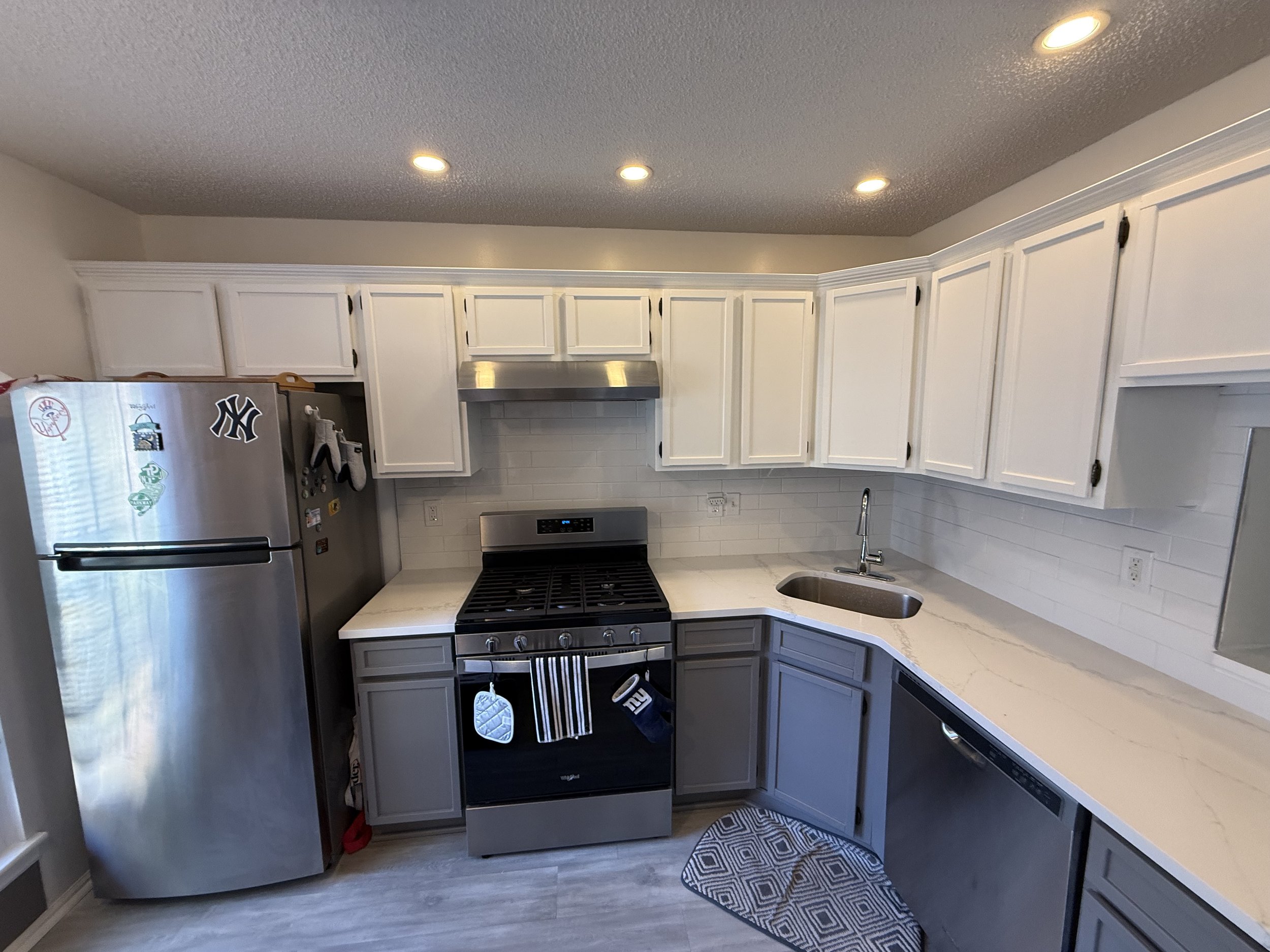 Modern kitchen with white upper cabinets, gray lower cabinets, stainless steel appliances, marble countertop, white subway tile backsplash, and a small rug on the floor.