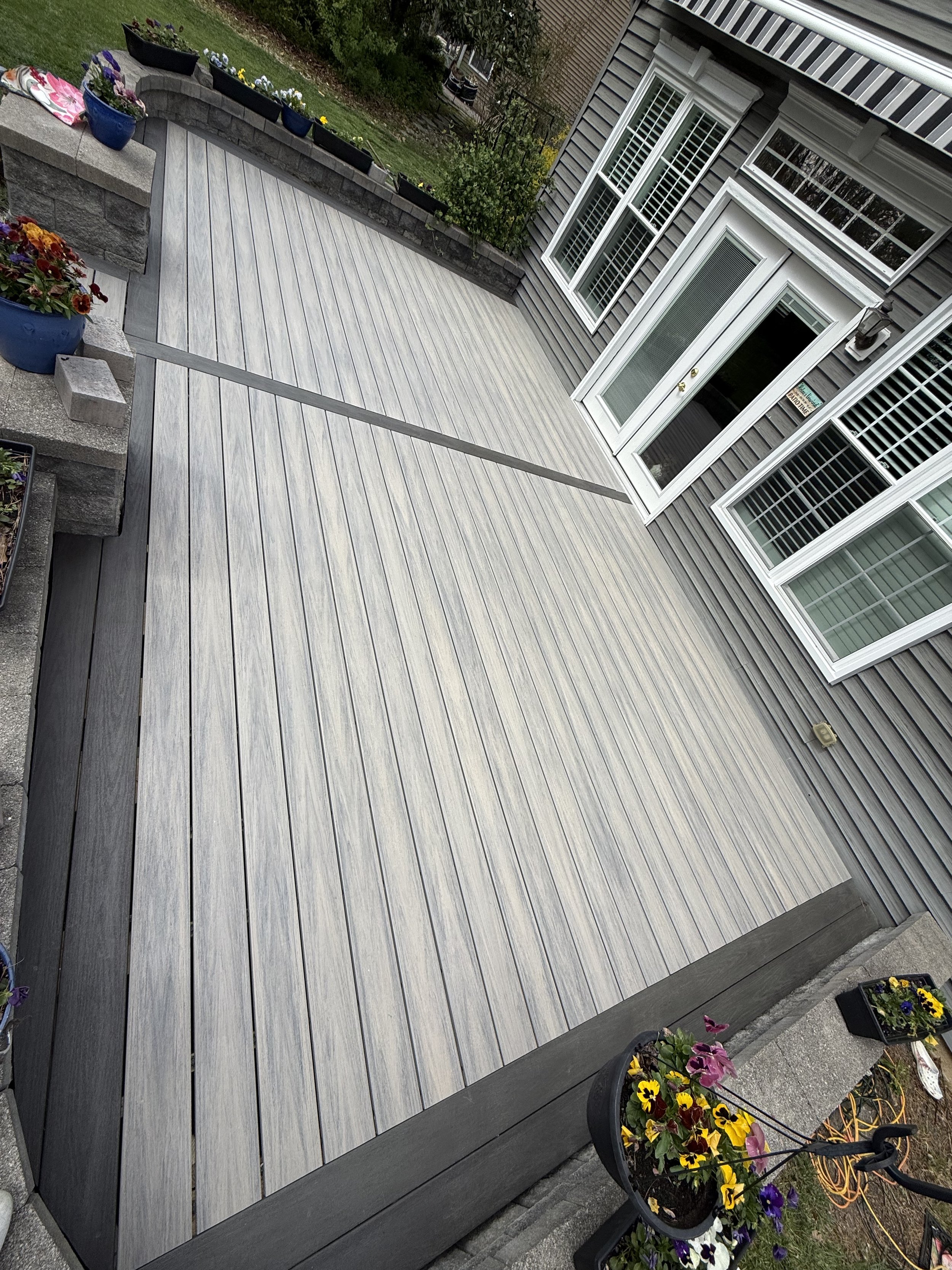 Newly built outdoor wooden deck with planters and flowers surrounding it, attached to a house with large Windows and a sliding glass door.