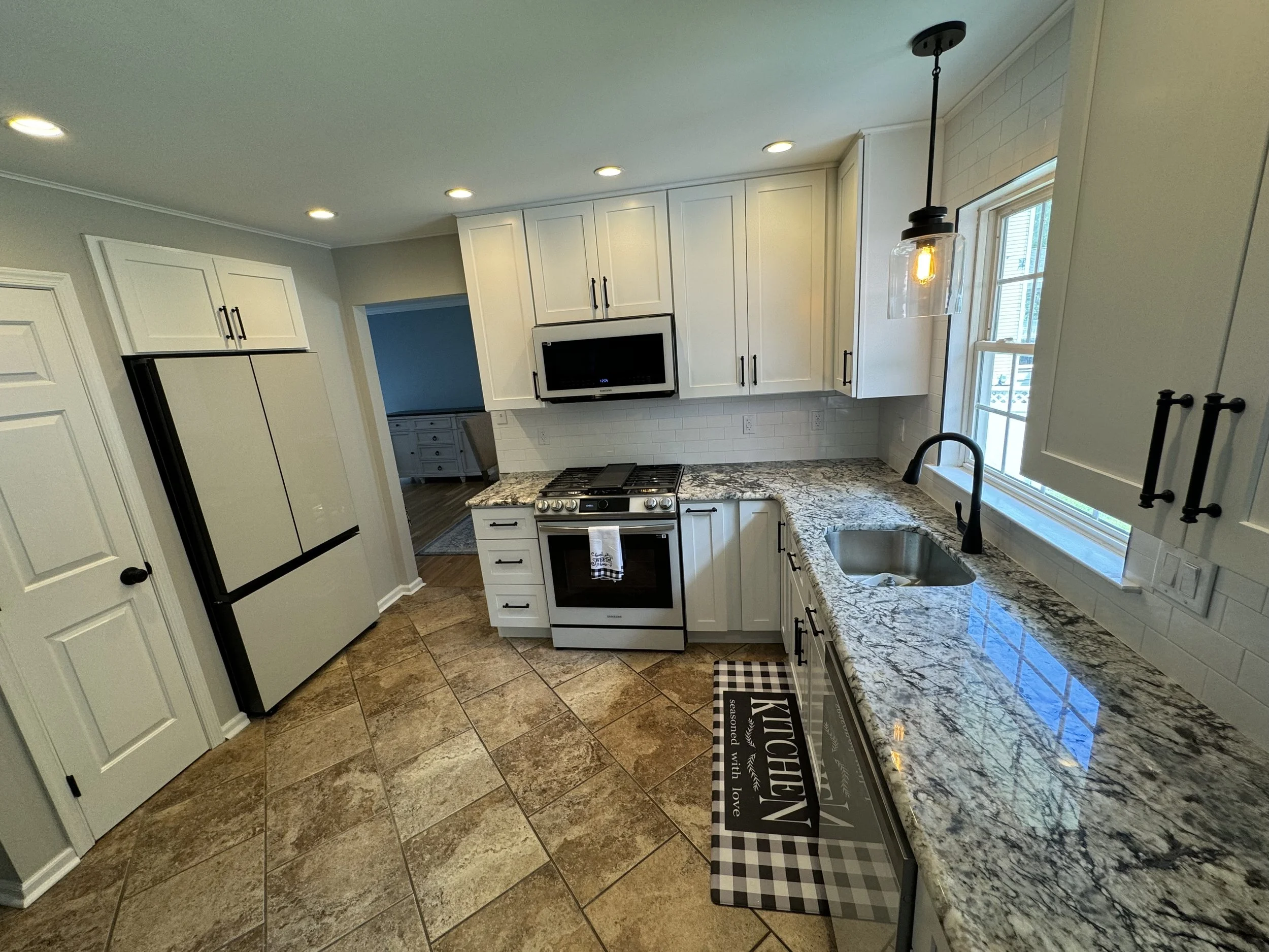 Modern kitchen with white cabinets, granite countertops, a stainless steel stove, a microwave above the stove, a black faucet, a window, and a tiled floor.