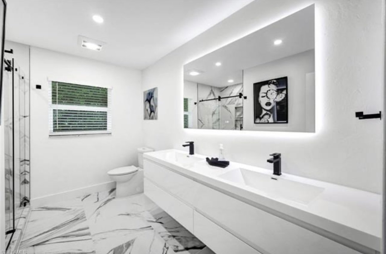 Modern white bathroom with dual black faucets, large mirror, artwork, marble floor, window with blinds, and shower enclosure.