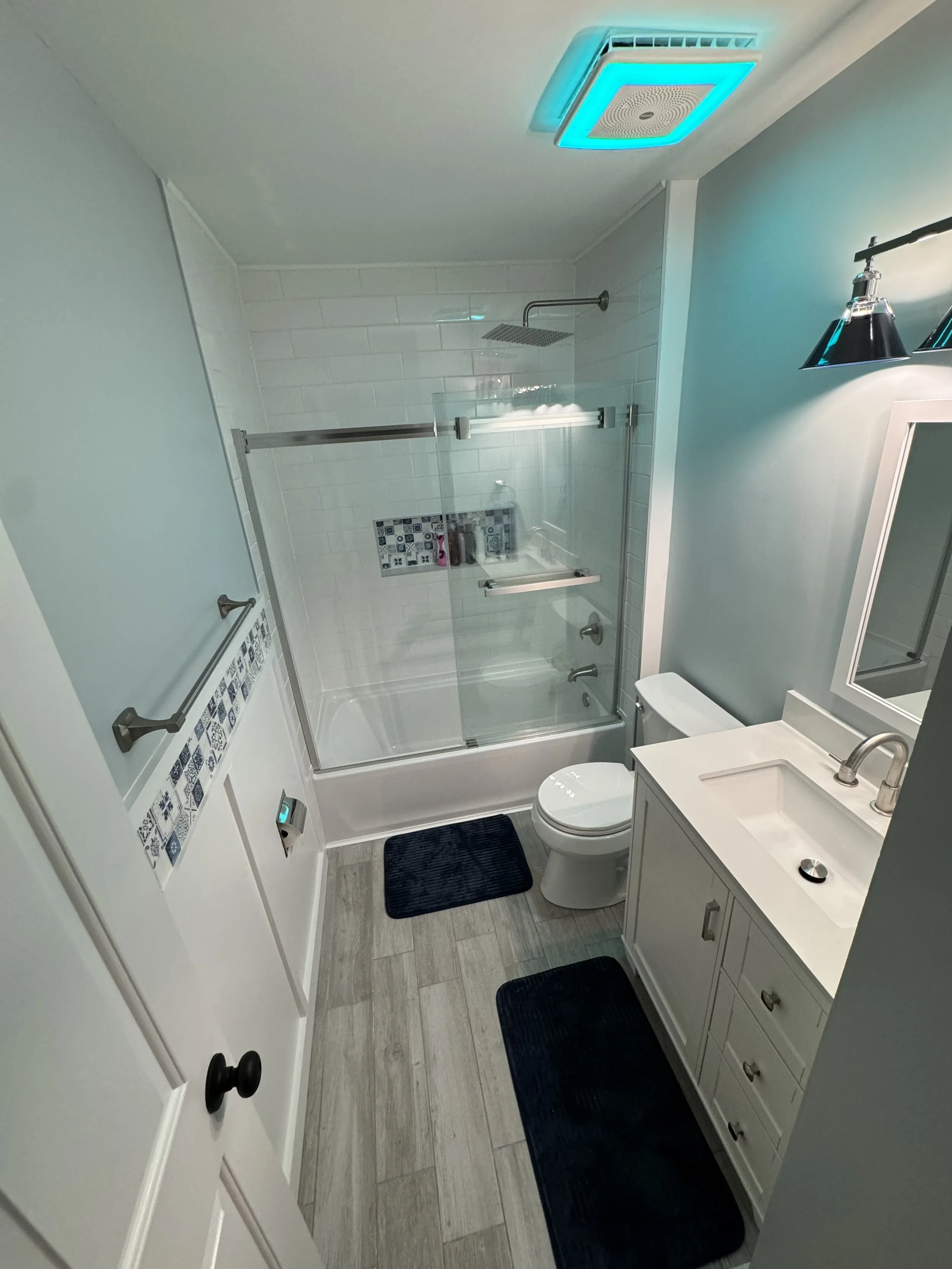A small modern bathroom with a shower stall, toilet, and white vanity with a sink and mirror. The shower has a sliding glass door, subway tile, and a built-in shelf. The ceiling has a turquoise vent. Two dark mats are on the light wood floor.