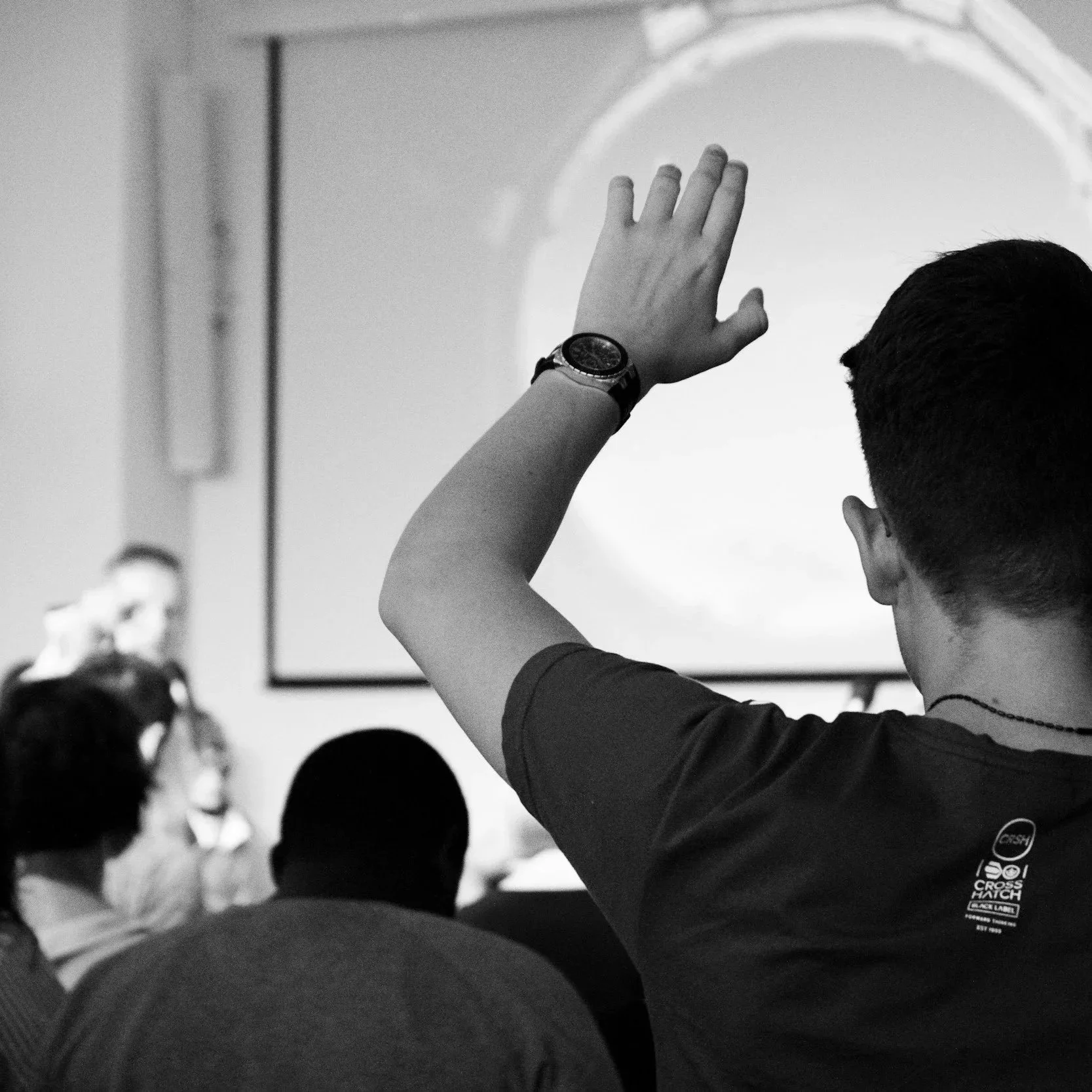 A person wearing a watch is raising their hand in front of a projection screen with an audience in a classroom or conference setting.