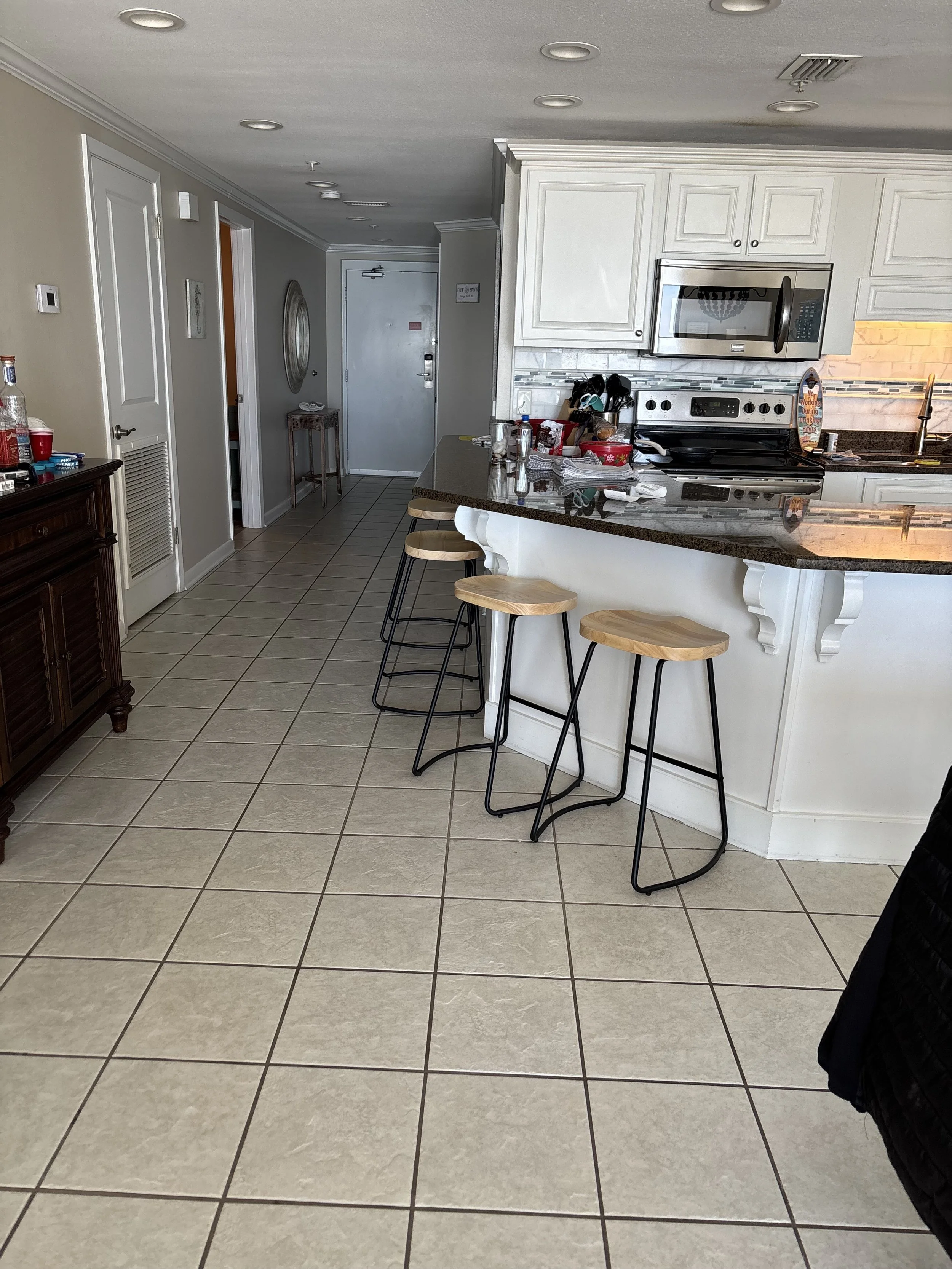 Interior view of a kitchen with white cabinets, a gray countertop, and a tiled floor. There are three wooden bar stools at the counter, and various kitchen items like bottles, utensils, and a tray on the counter. A microwave oven is built into the cabinets, and there's a sink with a window above it. In the background, there's a door and some decorative elements on the walls.