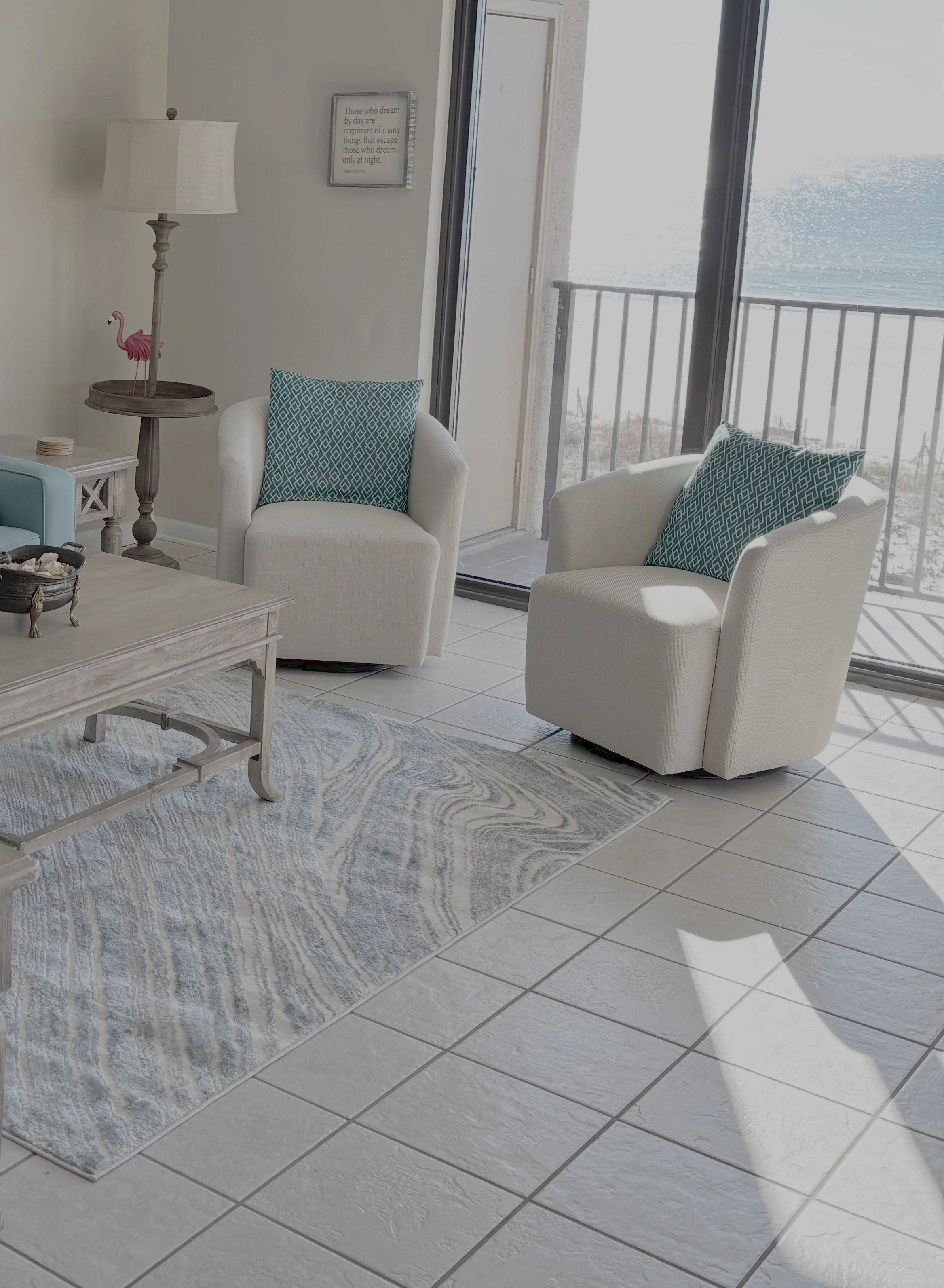 A bright living room corner with two white armchairs with blue patterned pillows, a wooden coffee table, a standing lamp with a pink flamingo decoration, a white side table, and a sliding glass door leading to an outdoor balcony.