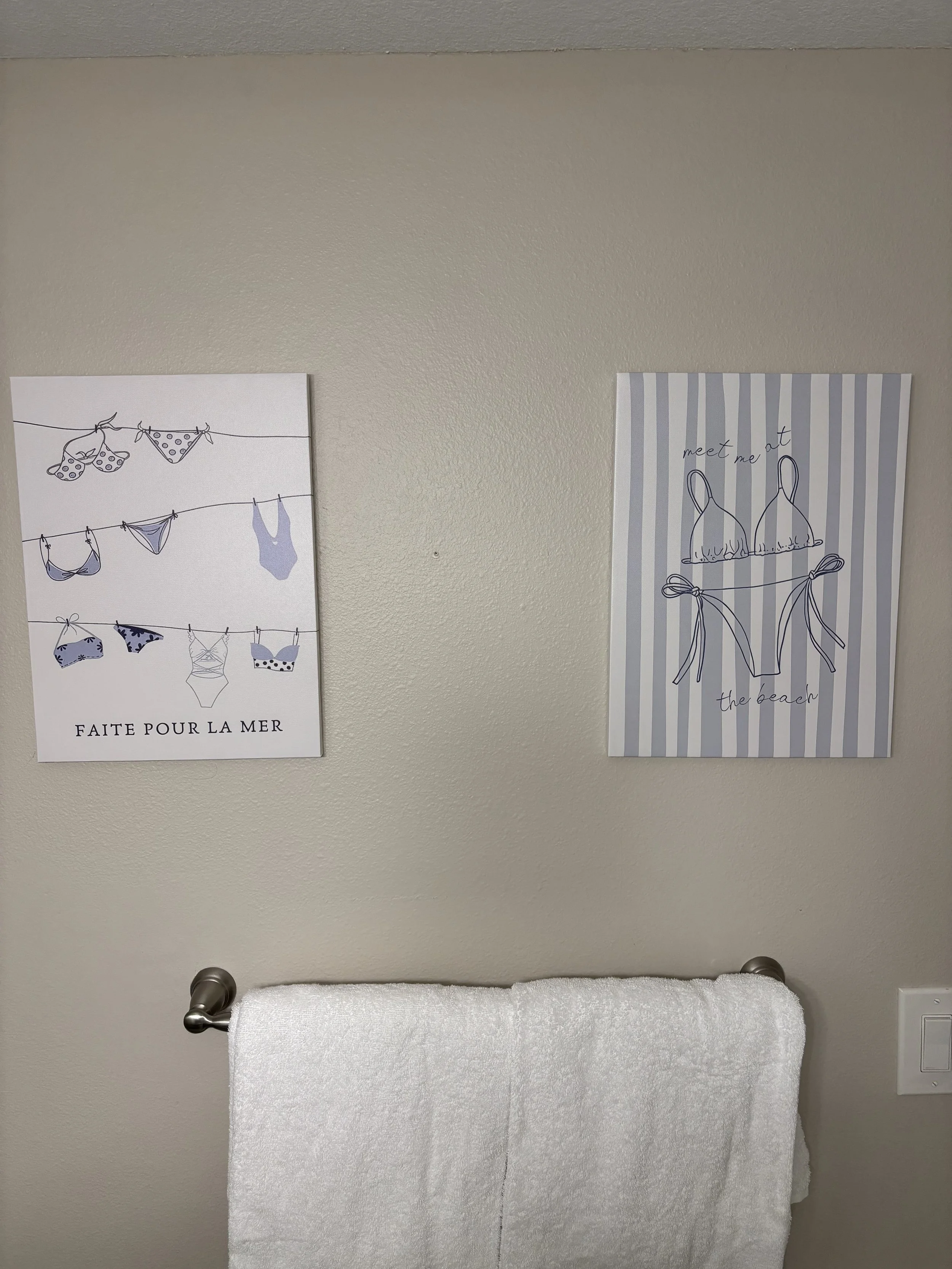 A white towel hanging on a towel bar in front of a beige wall with two decorative posters about swimwear and ocean themes.