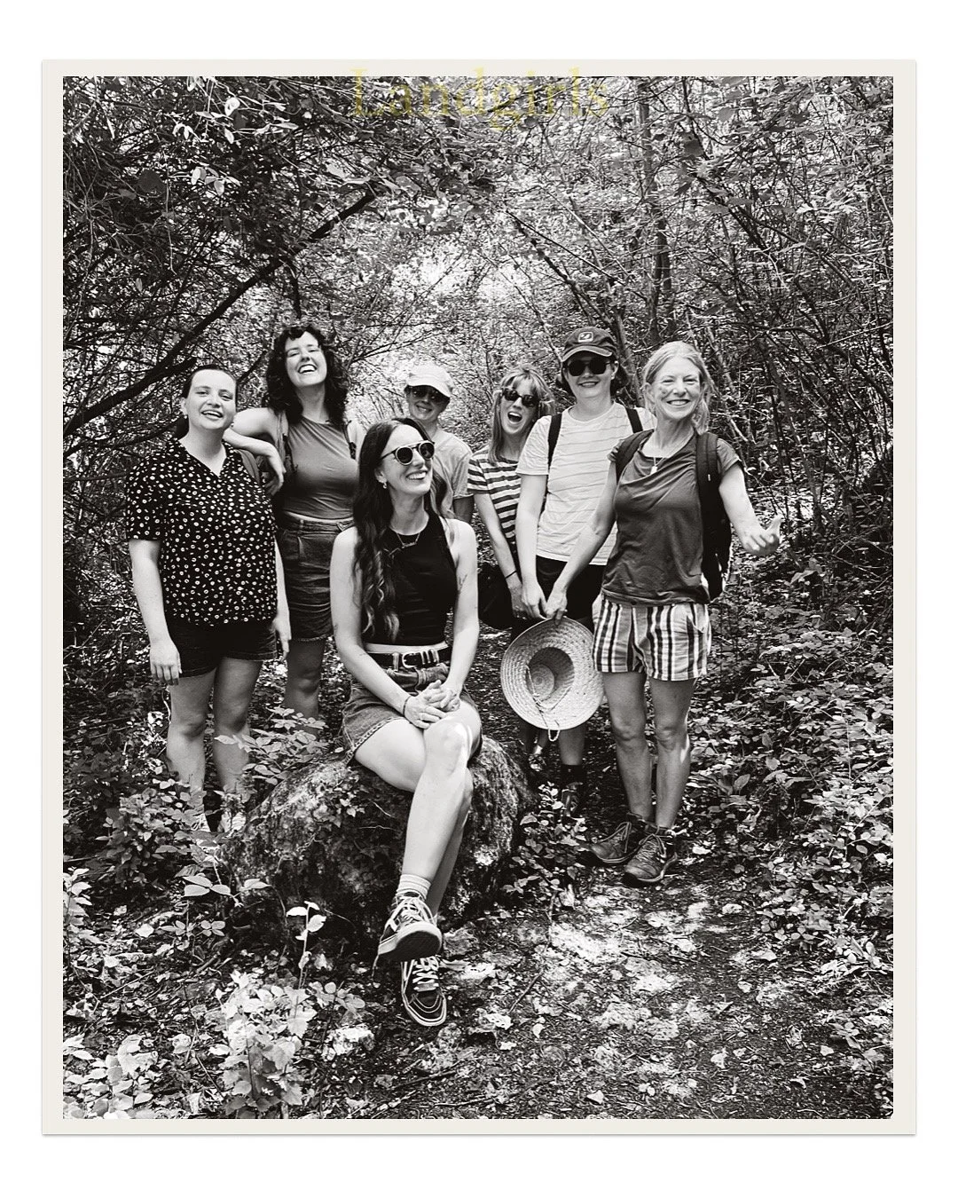 A group of eight women hiking in a wooded area, some wearing sunglasses and hats, smiling and posing for the photo.