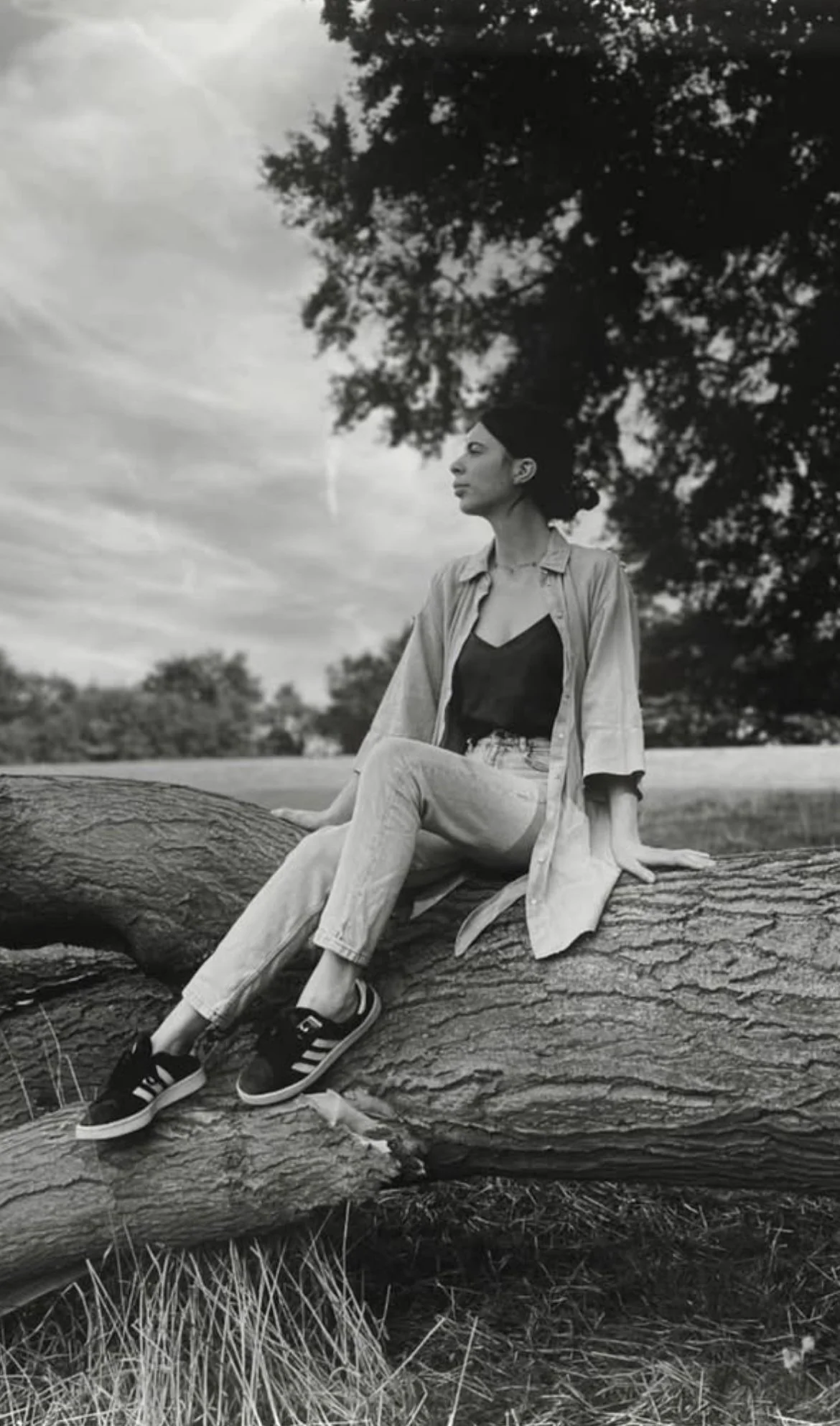 A woman sitting on a large fallen tree trunk outdoors, with her eyes closed and head turned to the side, surrounded by trees and cloudy sky.