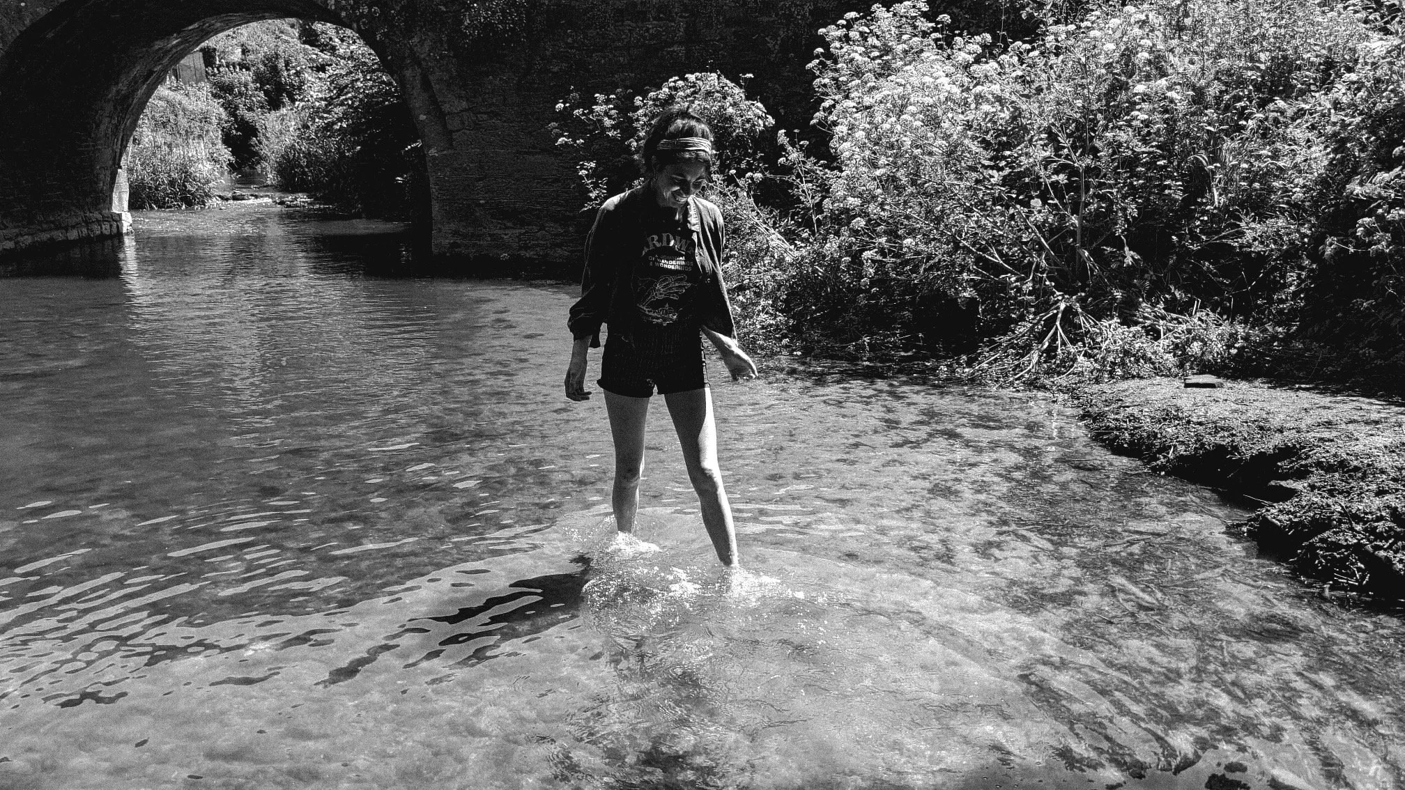 A person wading in a shallow river, under a stone bridge, surrounded by lush trees and bushes.