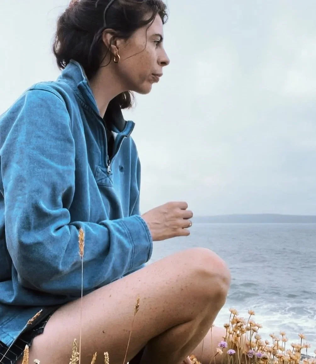 A woman sitting outdoors near the ocean, wearing a blue jacket, with brown hair and gold hoop earrings, surrounded by wildflowers, gazing at the sea.