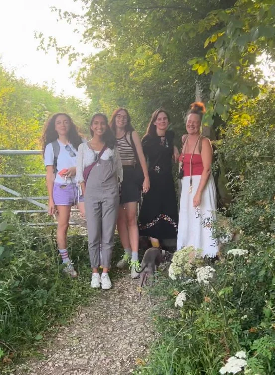 Group of five women standing on a nature trail surrounded by green foliage and trees with white flowers in the foreground.