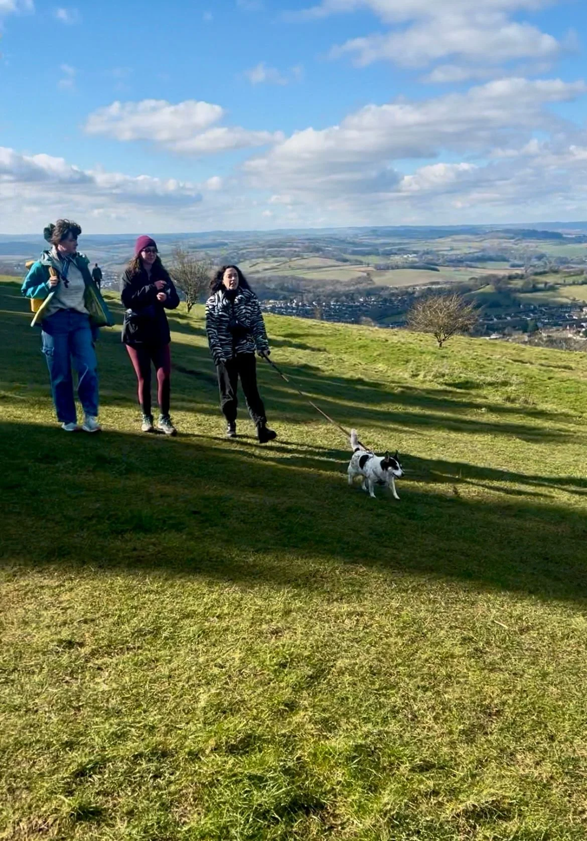 Three women and a man walking a dog on a grassy hill with a countryside view under a blue sky with clouds.