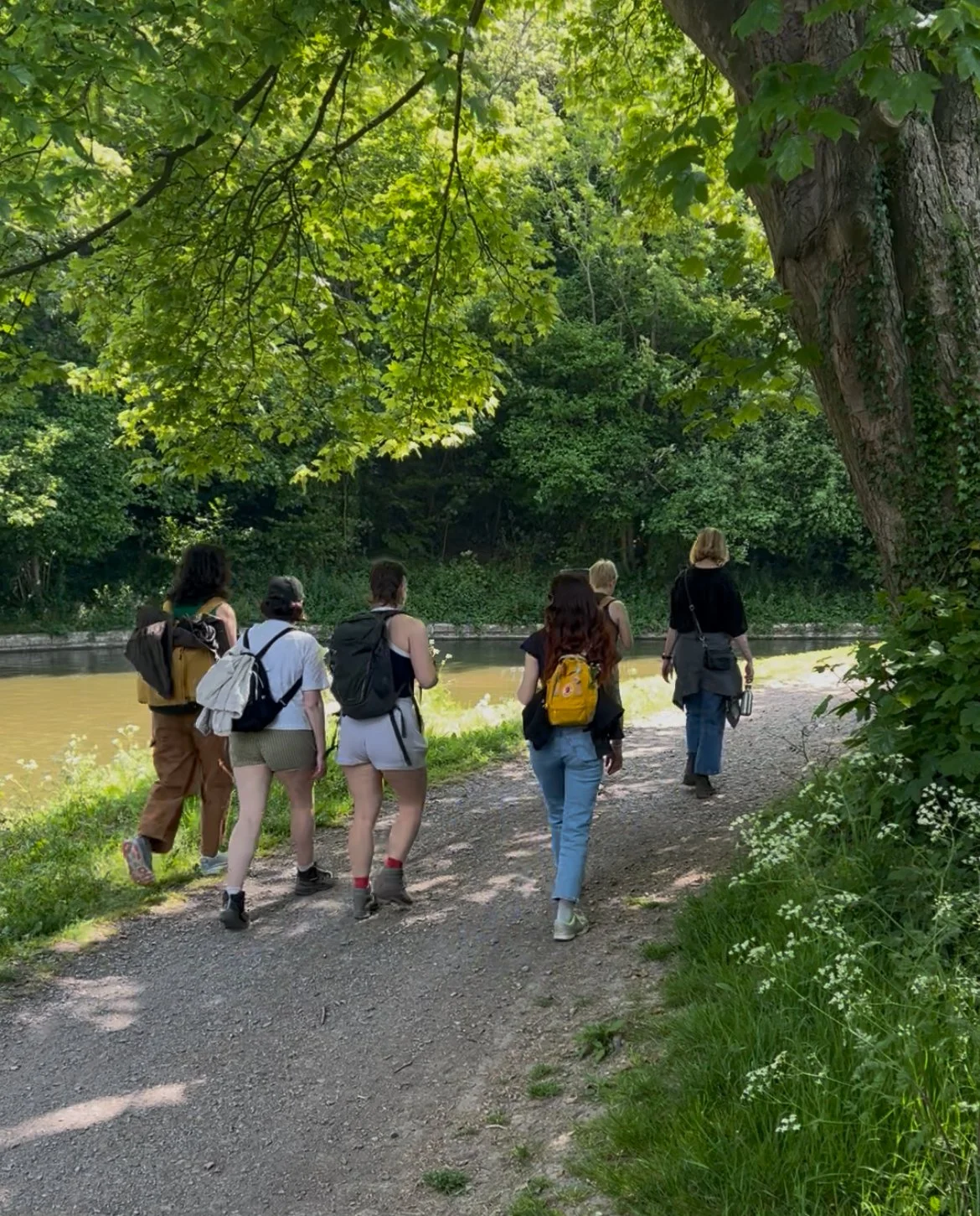Group of six people walking along a dirt path beside a river, surrounded by lush green trees and foliage.
