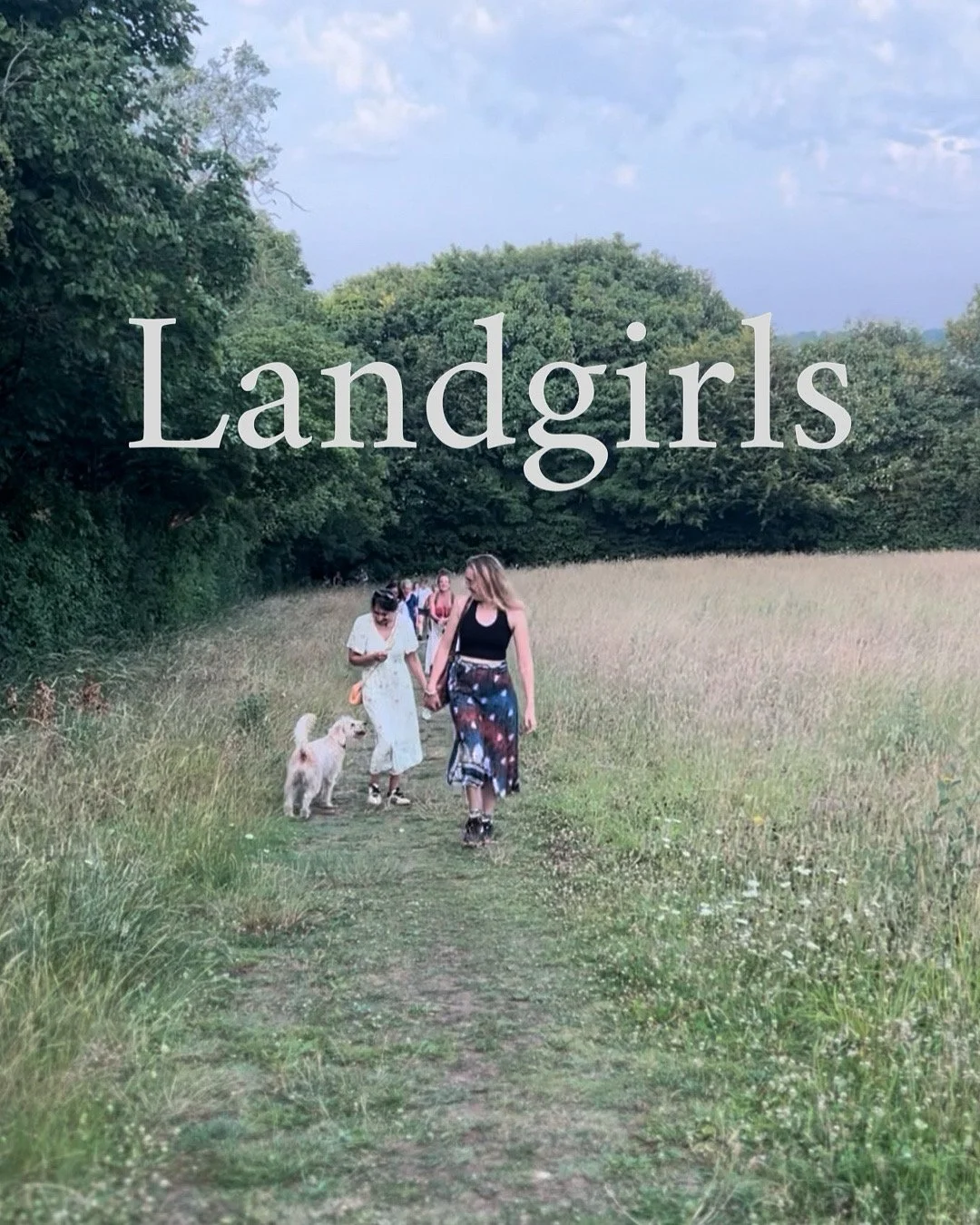 A group of women, some holding hands and walking a dog, walk along a grassy trail through a lush, green field with trees in the background, under a partly cloudy sky.