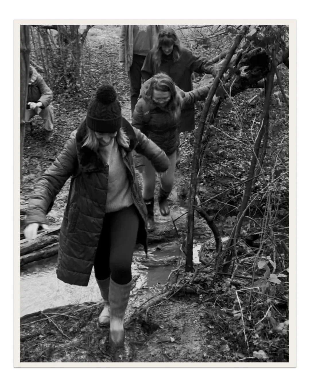 People crossing a small stream in a wooded area, some wearing warm clothing and boots.