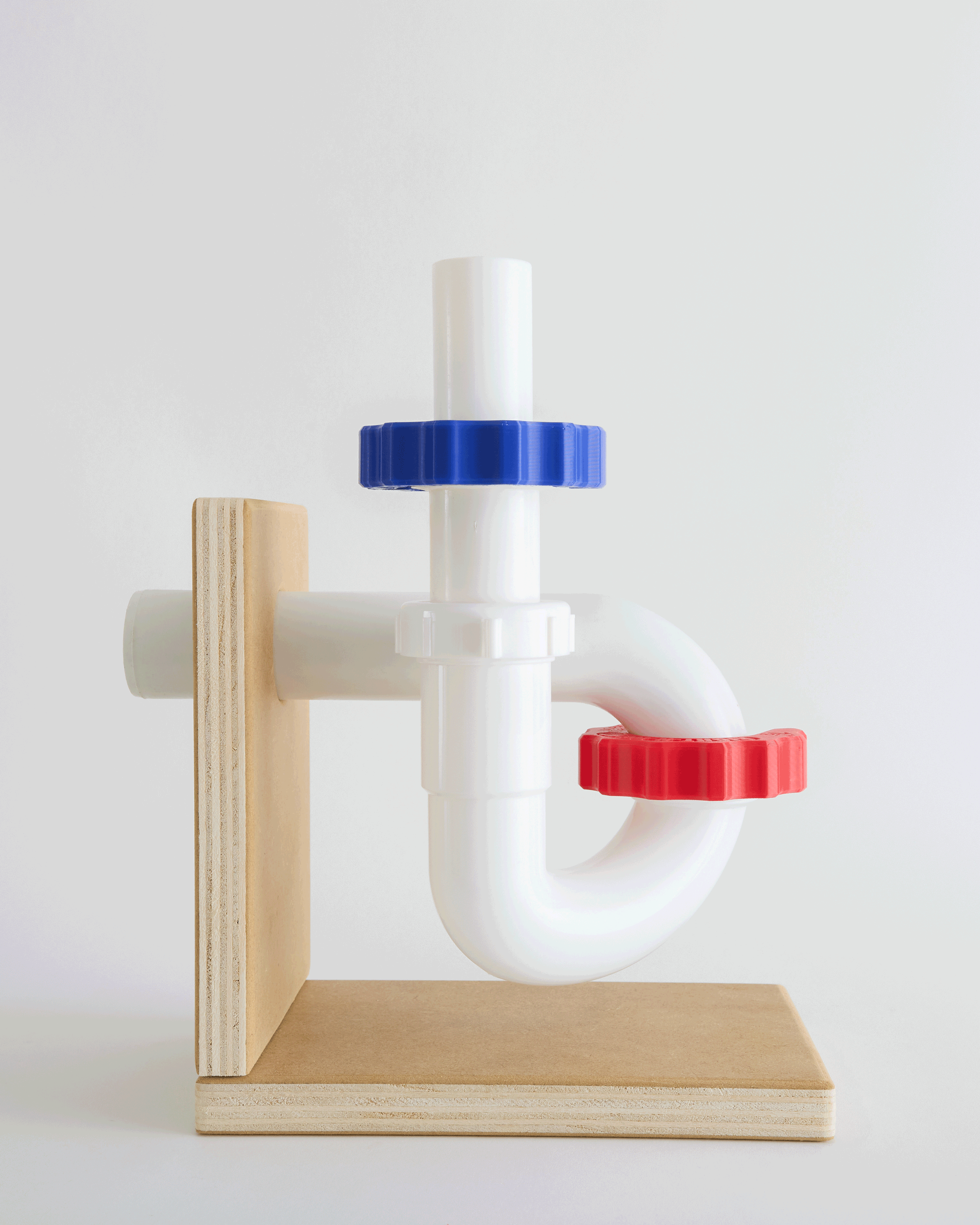A colorful pipe sculpture with red, blue, and white pipe fittings mounted on a wooden stand.