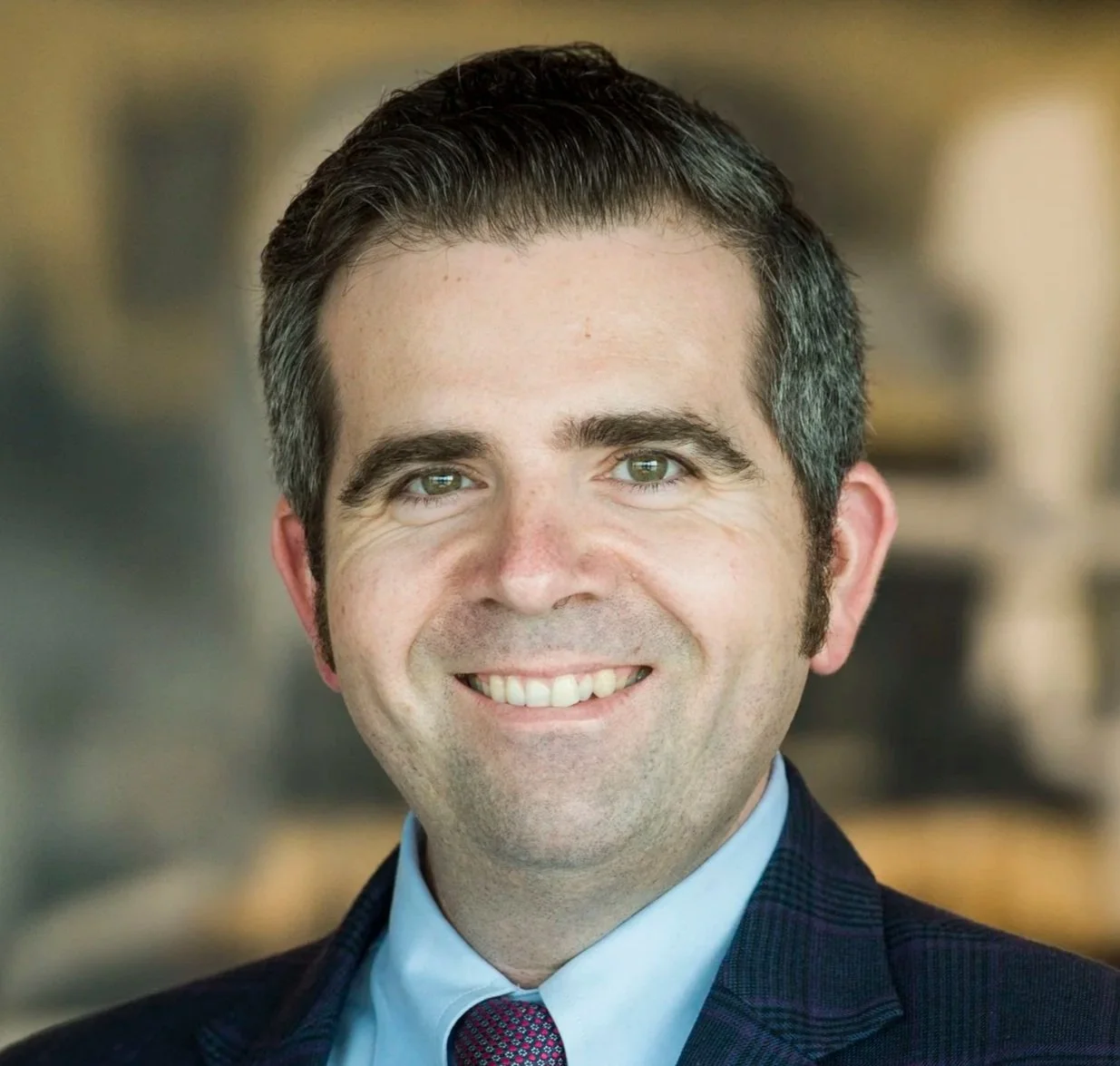 Gene Tunny =  A smiling man with dark hair, light shirt, and navy blazer in an office setting.