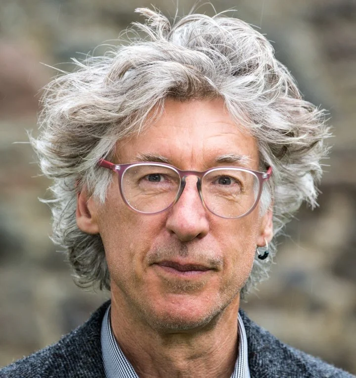 Alex Coram = A middle-aged man with grey hair, glasses, and an earring, wearing a grey jacket and collared shirt, looking into the camera with a neutral expression.