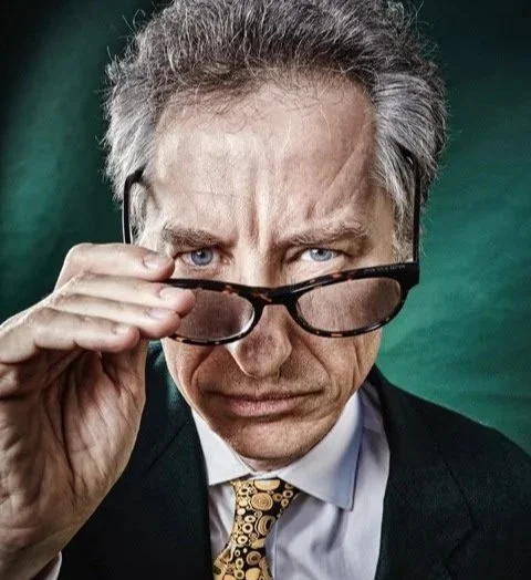 Nicholas Gruen = a man with gray hair and blue eyes adjusting his glasses, wearing a dark suit, white shirt, and patterned tie, looking intently at the camera against a dark green background.