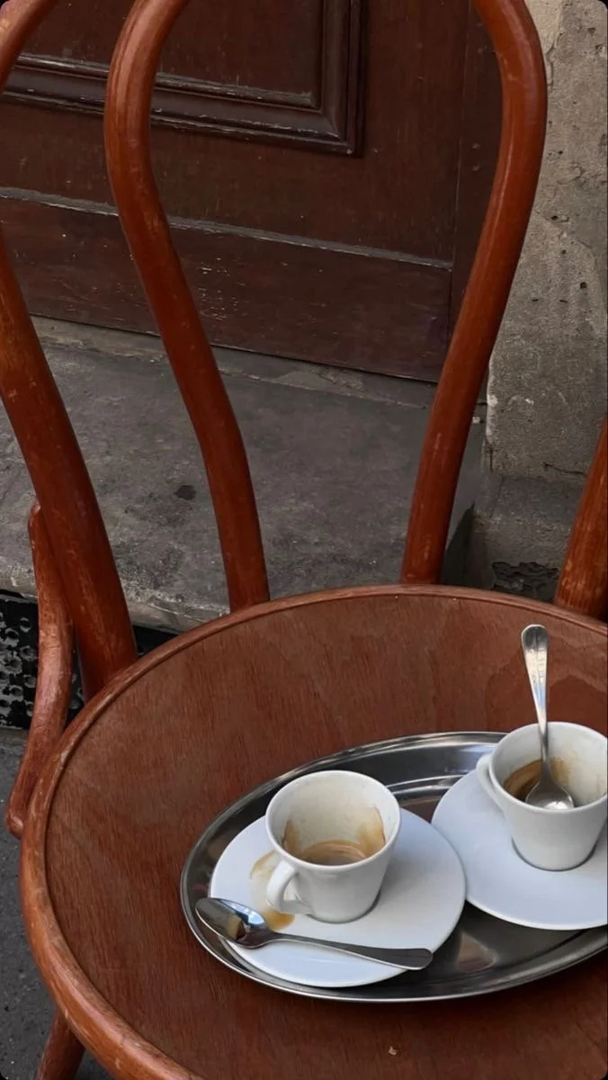Two empty white coffee cups with saucers and spoons on a metal tray on a wooden chair outside, in front of a brown door and stone wall.