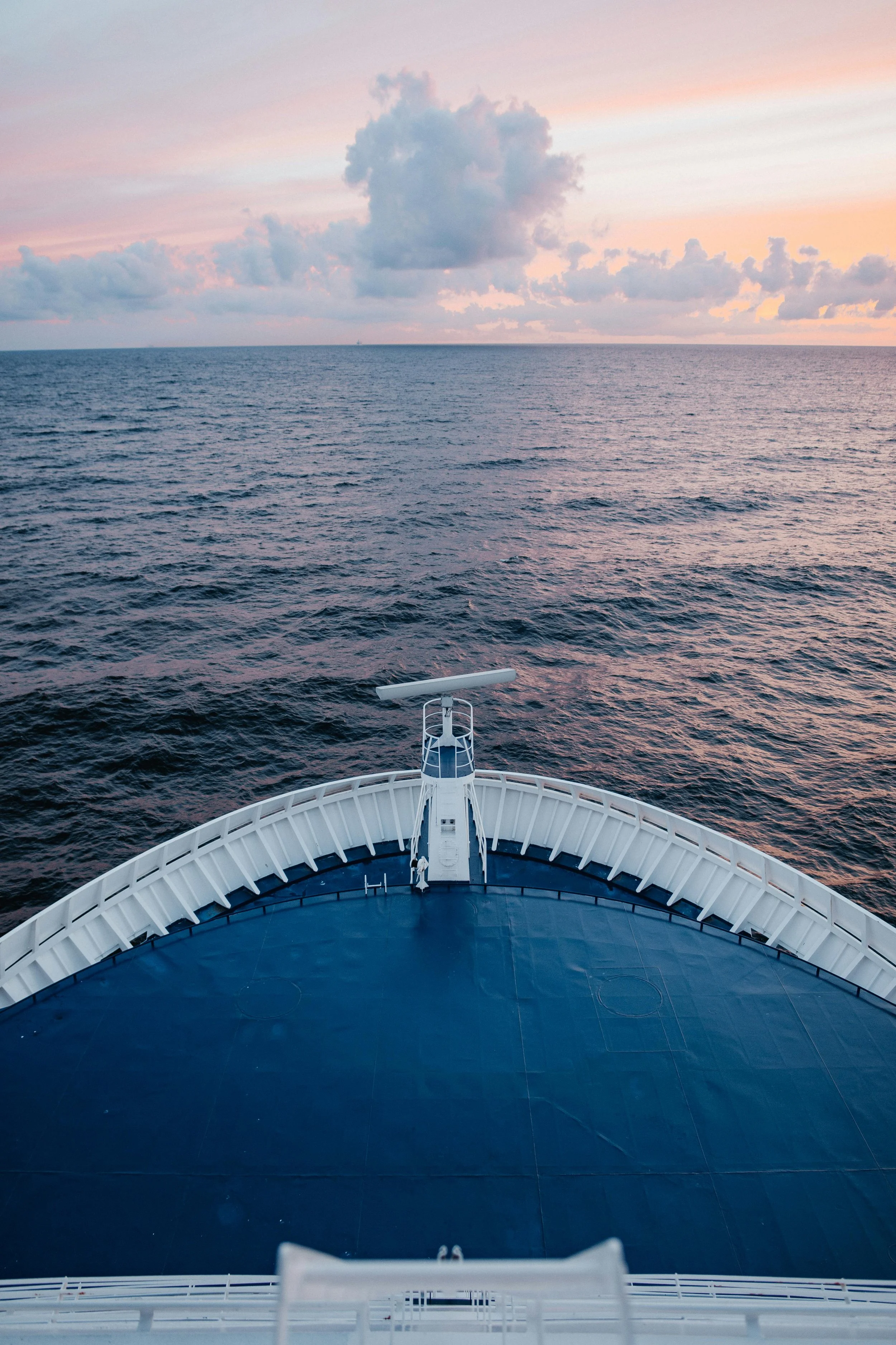 View from the front of a cruise ship during sunset, showing the ship's bow and the ocean with pink and blue sky, some clouds in the distance.