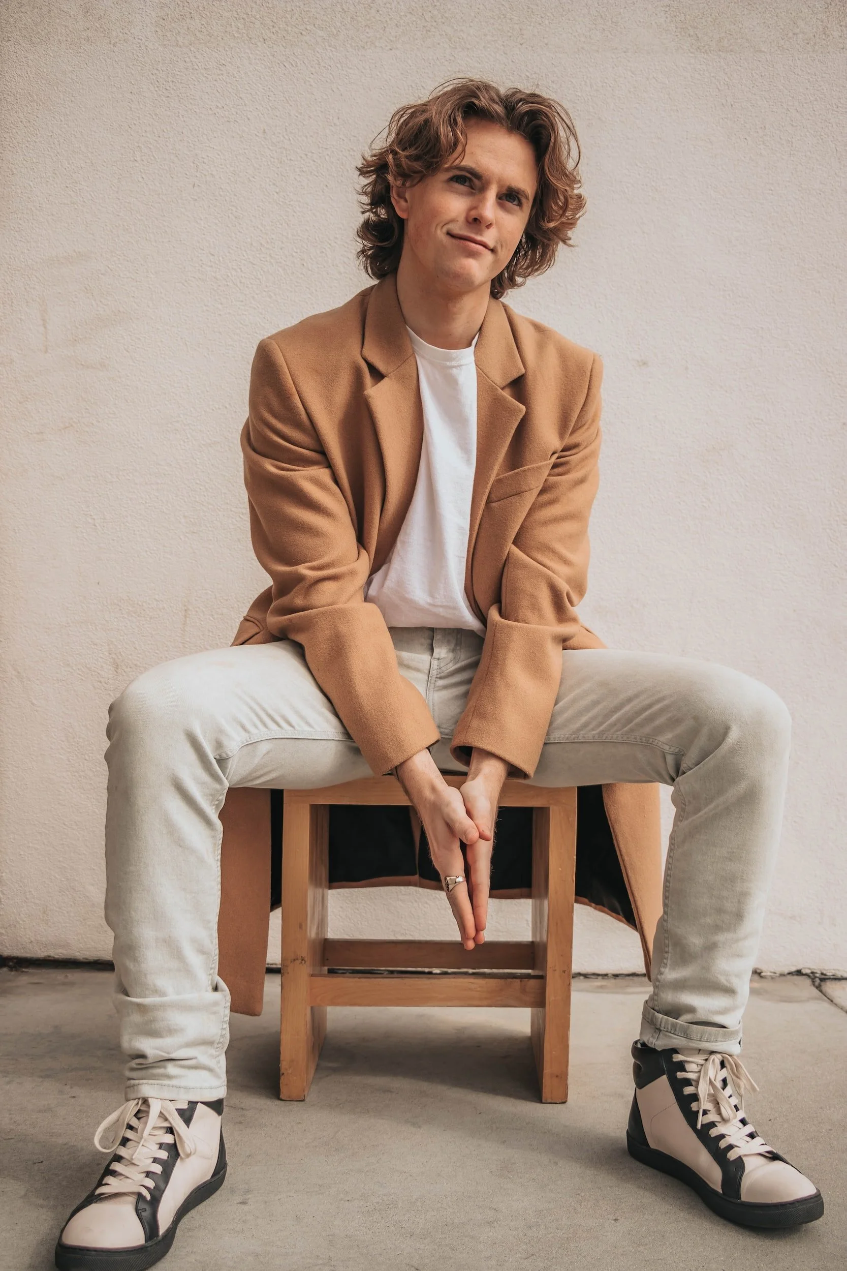 A young man with wavy, shoulder-length hair, sitting on a small wooden stool against a plain beige wall. He is wearing a camel-colored blazer, a white t-shirt, light-colored jeans, and black-and-white sneakers. He has a slight smile and is sitting with legs apart, leaning forward with his hands clasped between his knees.