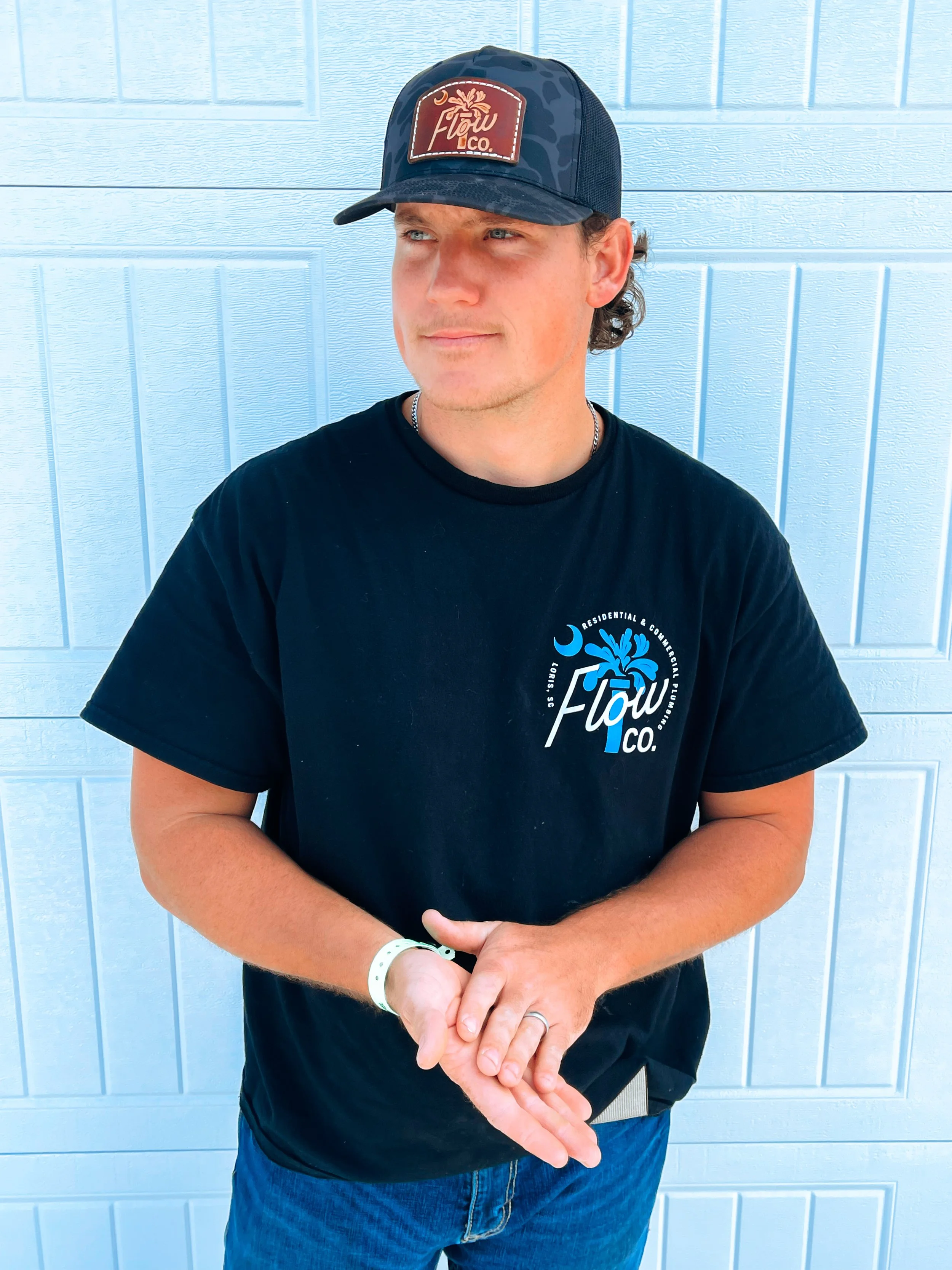 Young man wearing a black T-shirt with a logo, a black hat with a logo, standing in front of a light blue wall.