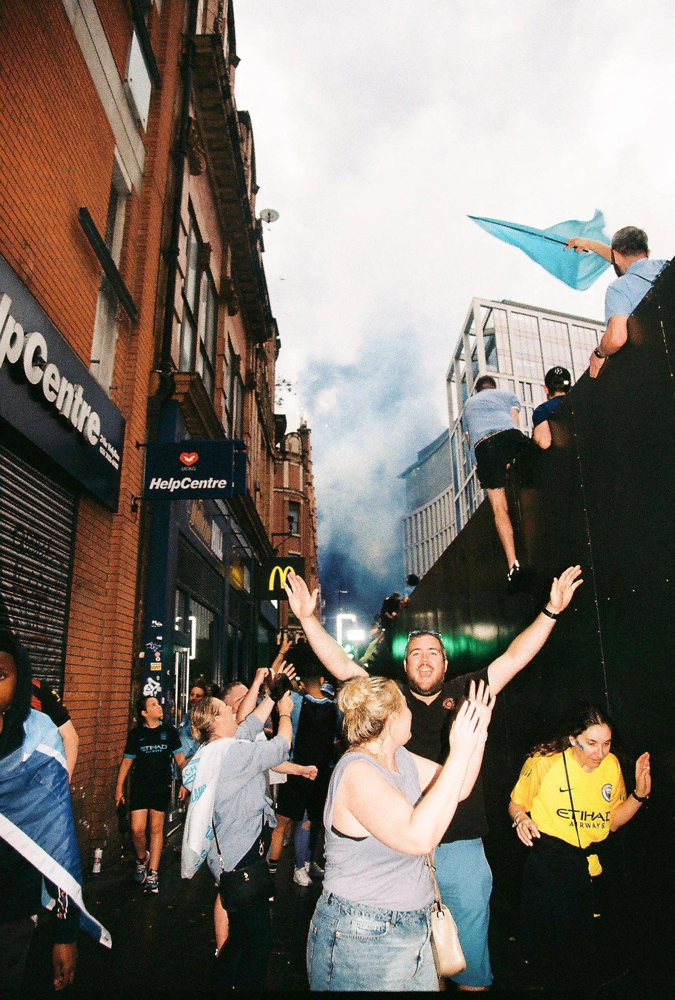 People celebrating on a city street after a fire or incident, with some climbing on a wall, smoke in the background, and bystanders cheering.