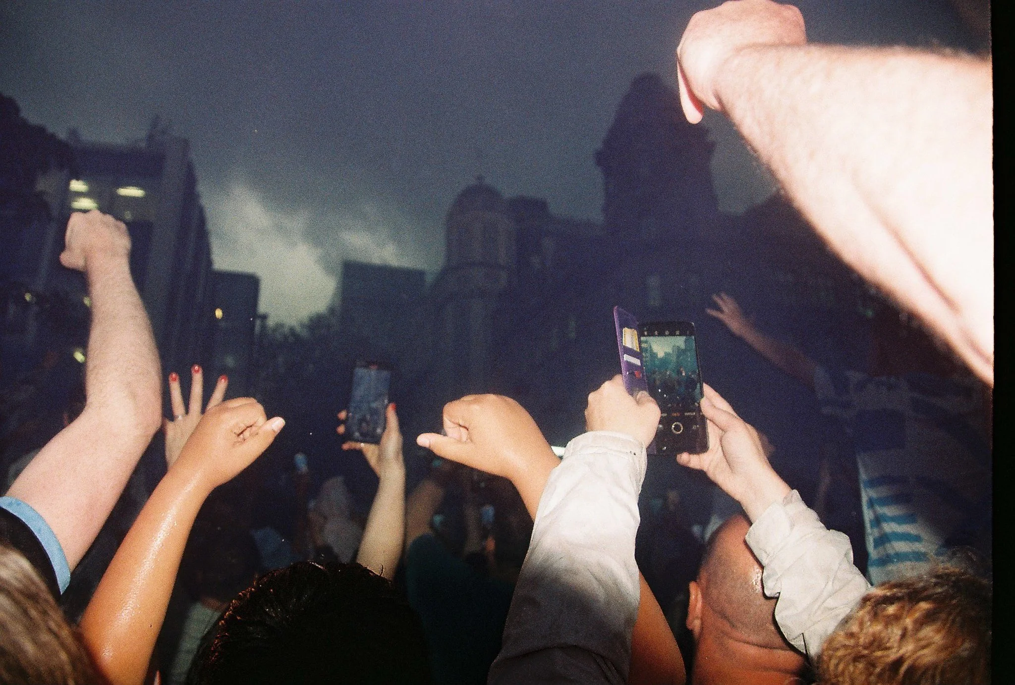 Concert crowd raising hands and taking photos with smartphones during a dark, outdoor concert at night.