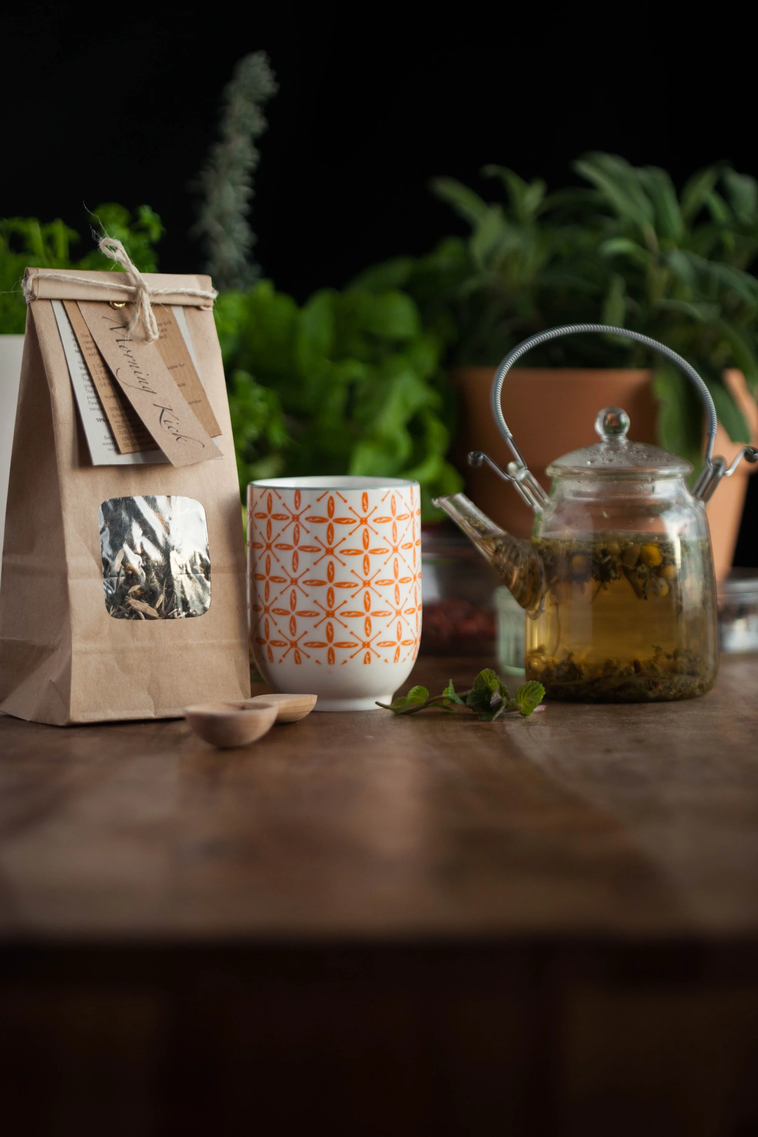 A brown paper bag of coffee, a white mug with an orange geometric pattern, a glass teapot with herbal tea and floating herbs, and a small wooden spoon on a wooden table with green plants in the background.