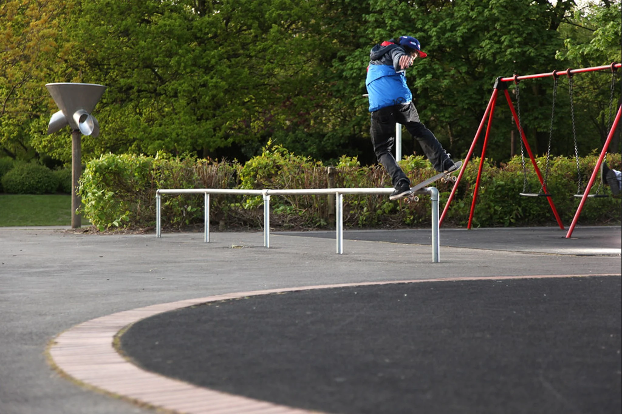 A person skateboarding over a metal rail at a park with green trees in the background and a swing set to the right.