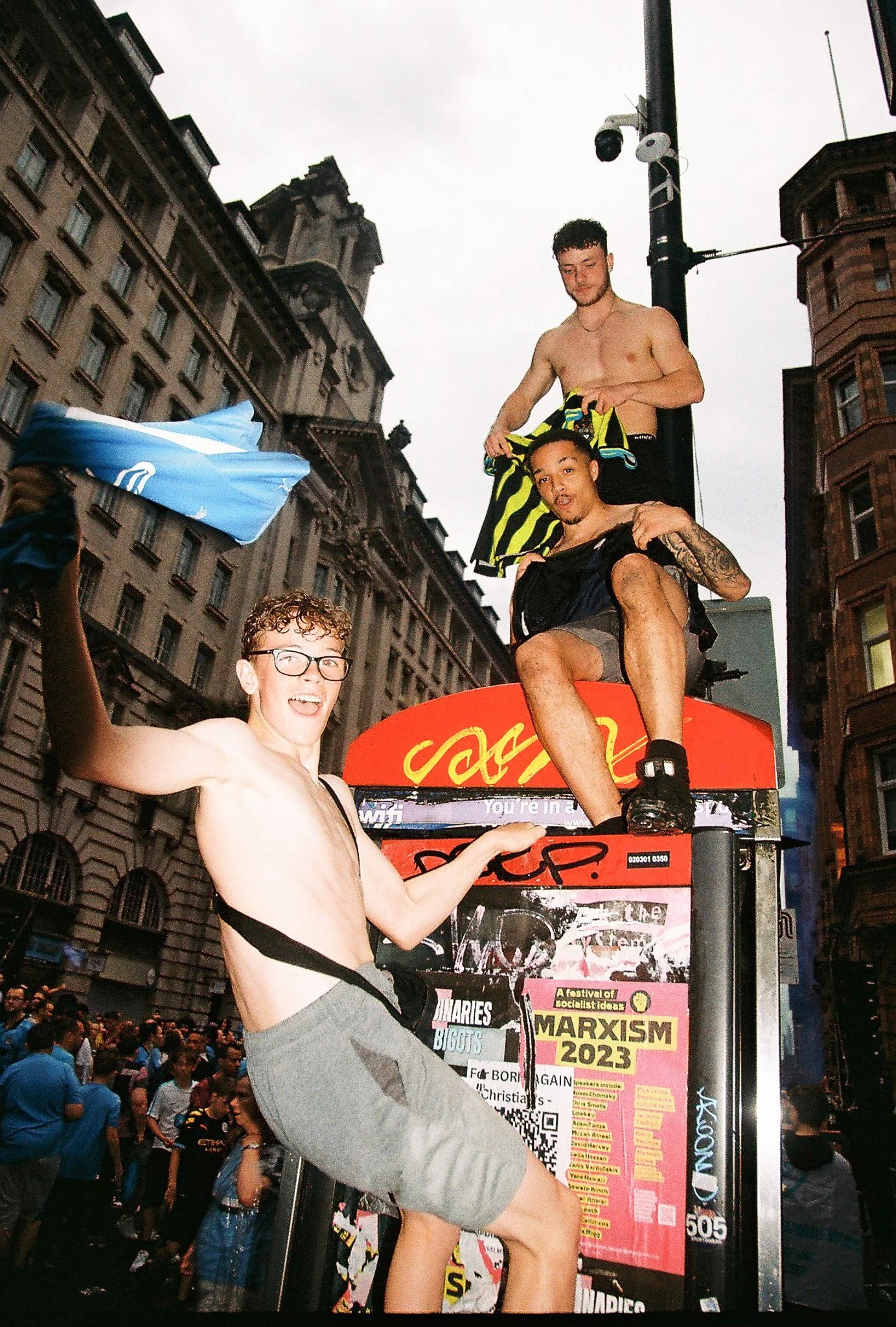 Three shirtless young men sitting and standing on top of a red bus in a city streets, surrounded by a crowd. One is holding a blue item, another is sitting with a tattoo on his arm, and the third is looking down.
