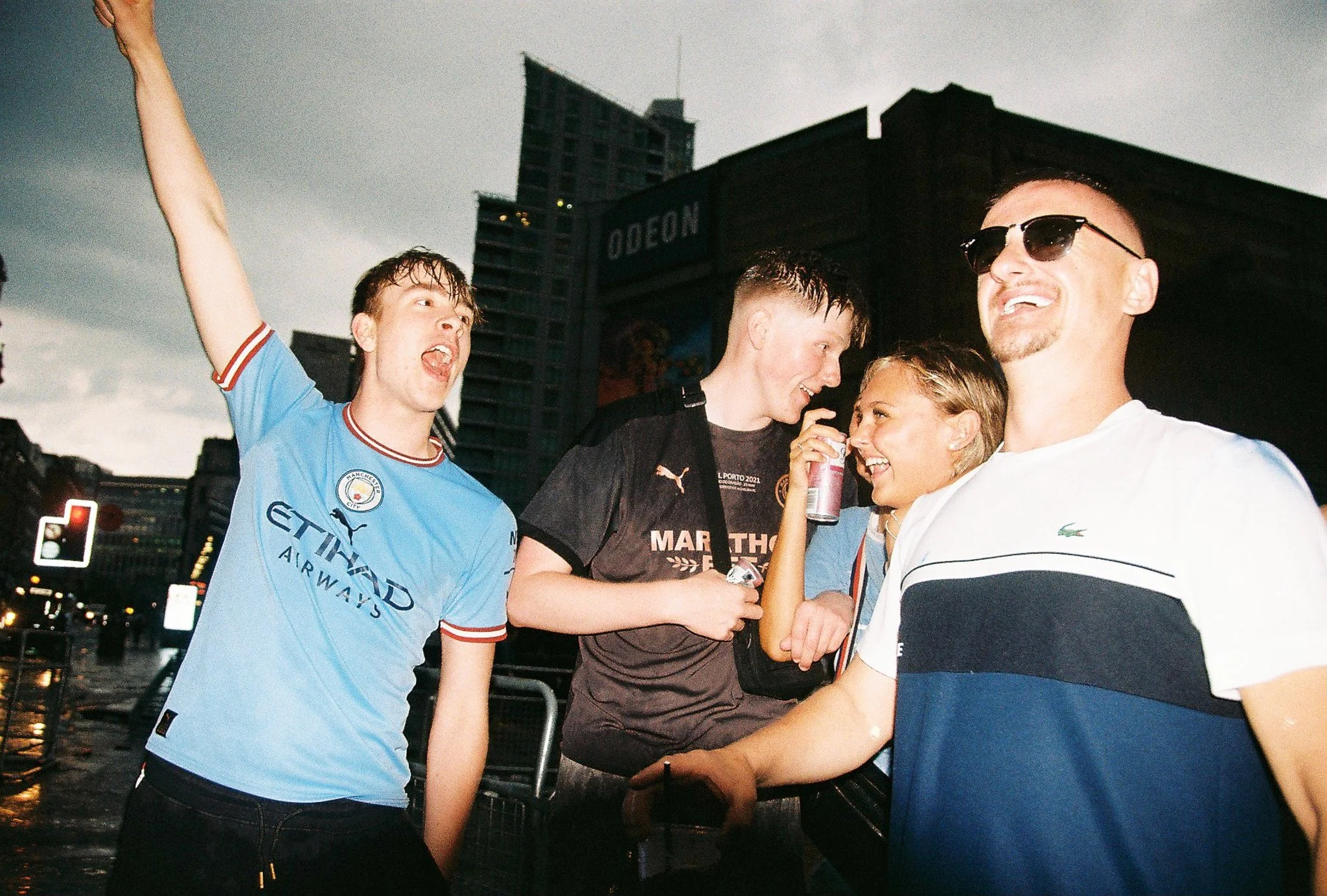 Group of young friends celebrating outdoors in an urban area during dusk, one person wearing a Manchester City football jersey, others smiling and holding drinks, with tall buildings and the ODEON sign in the background.