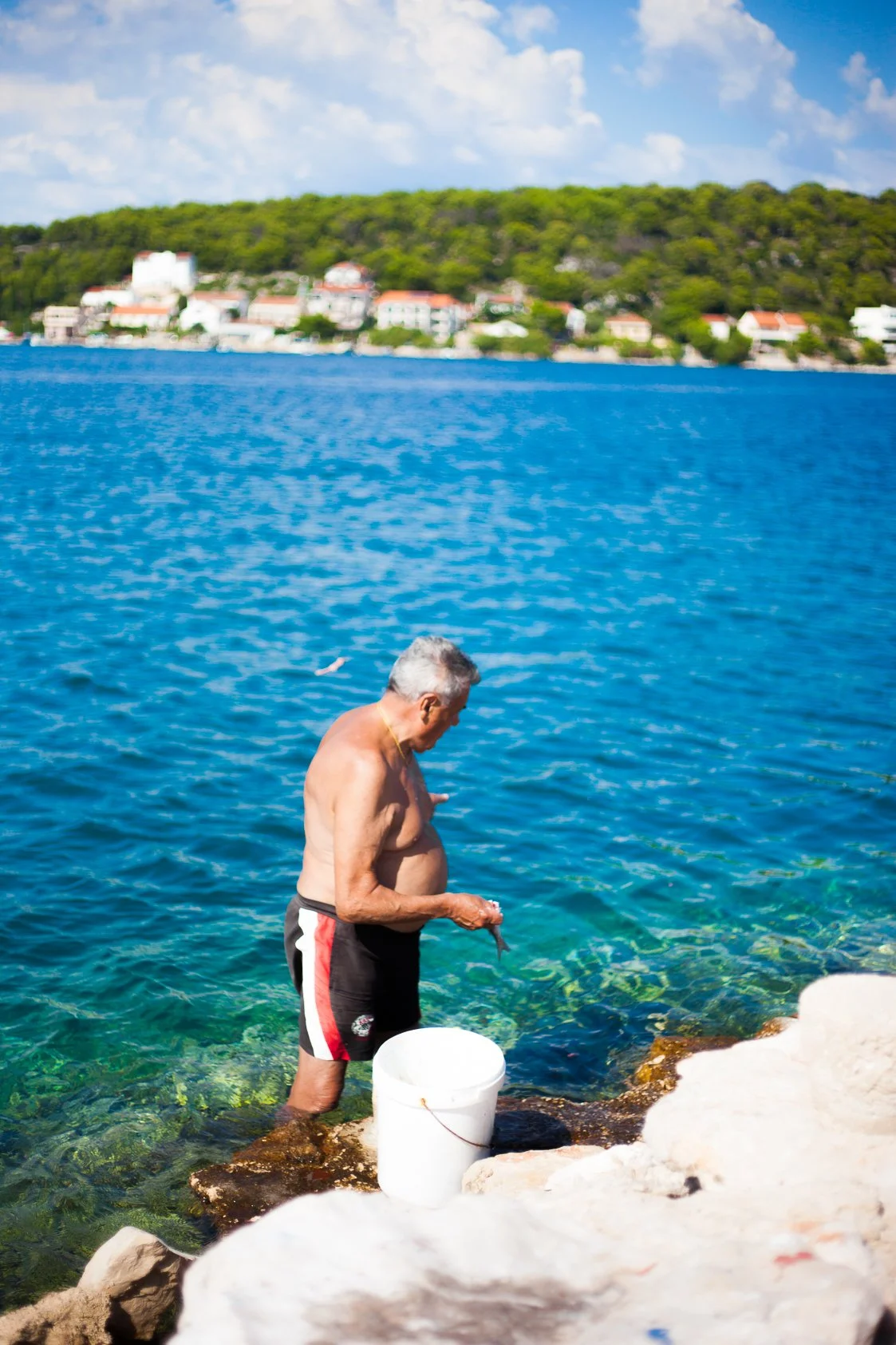 An elderly man fishing by the shore of a blue body of water, with a bucket nearby, and a hillside with houses in the background.