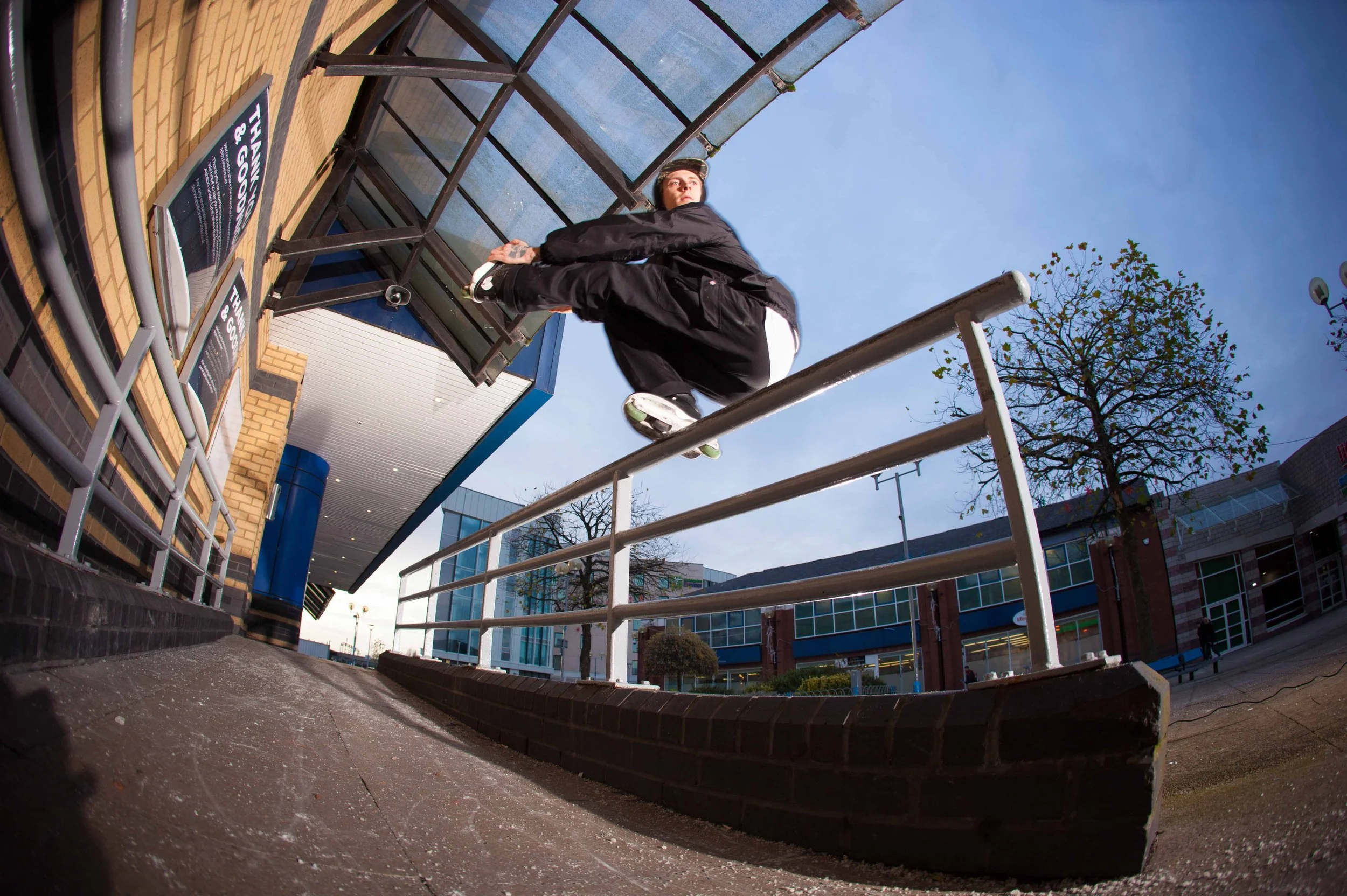 Teenager jumping over a metal railing outside a shopping mall or commercial building during daytime, with trees and modern architecture in the background.